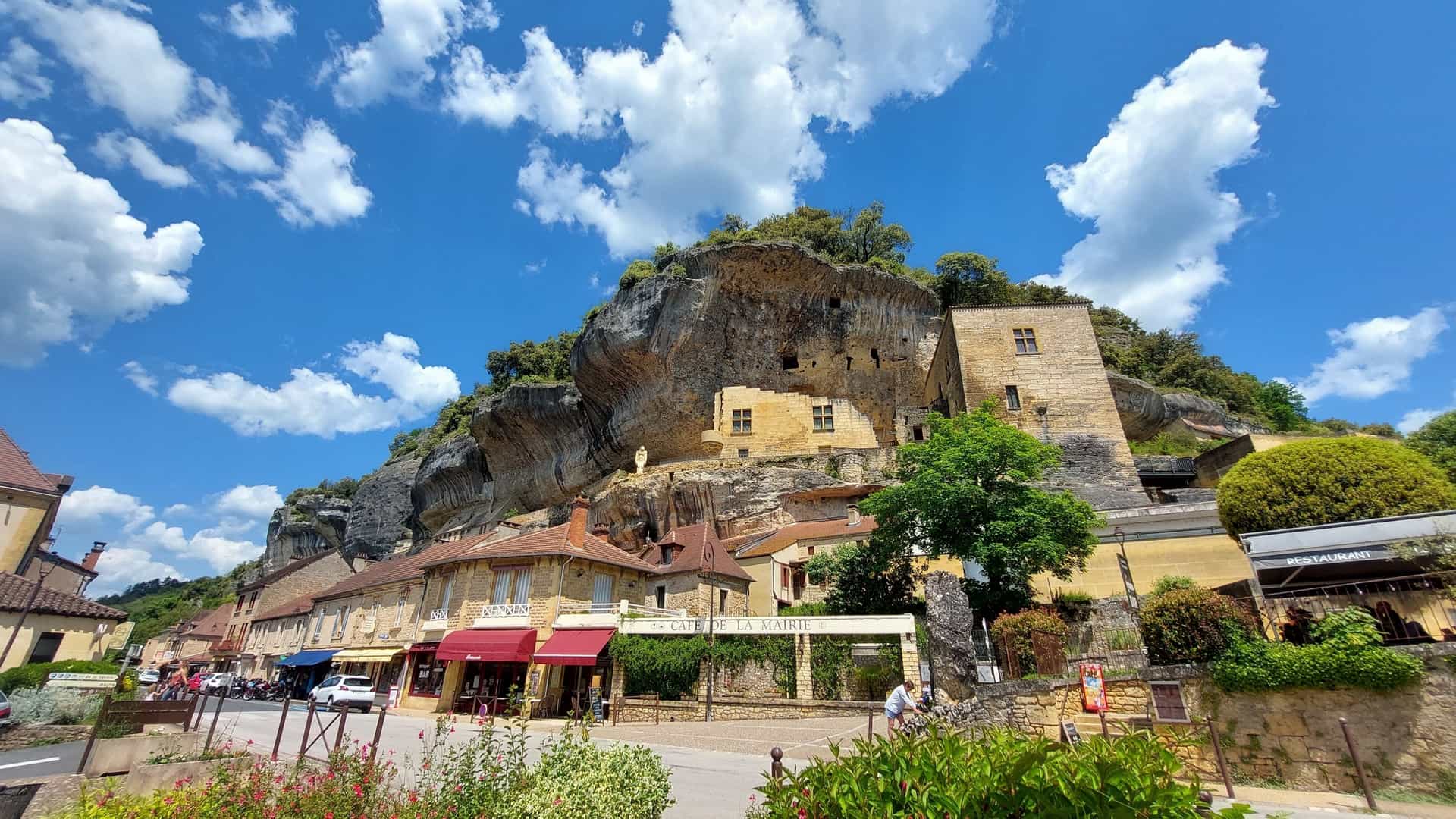 Medieval-style buildings in Les Eyzies-de-Tayac-Sireuil, France, are constructed into the base of a massive, overhanging limestone cliff under a bright blue, cloudy sky.
