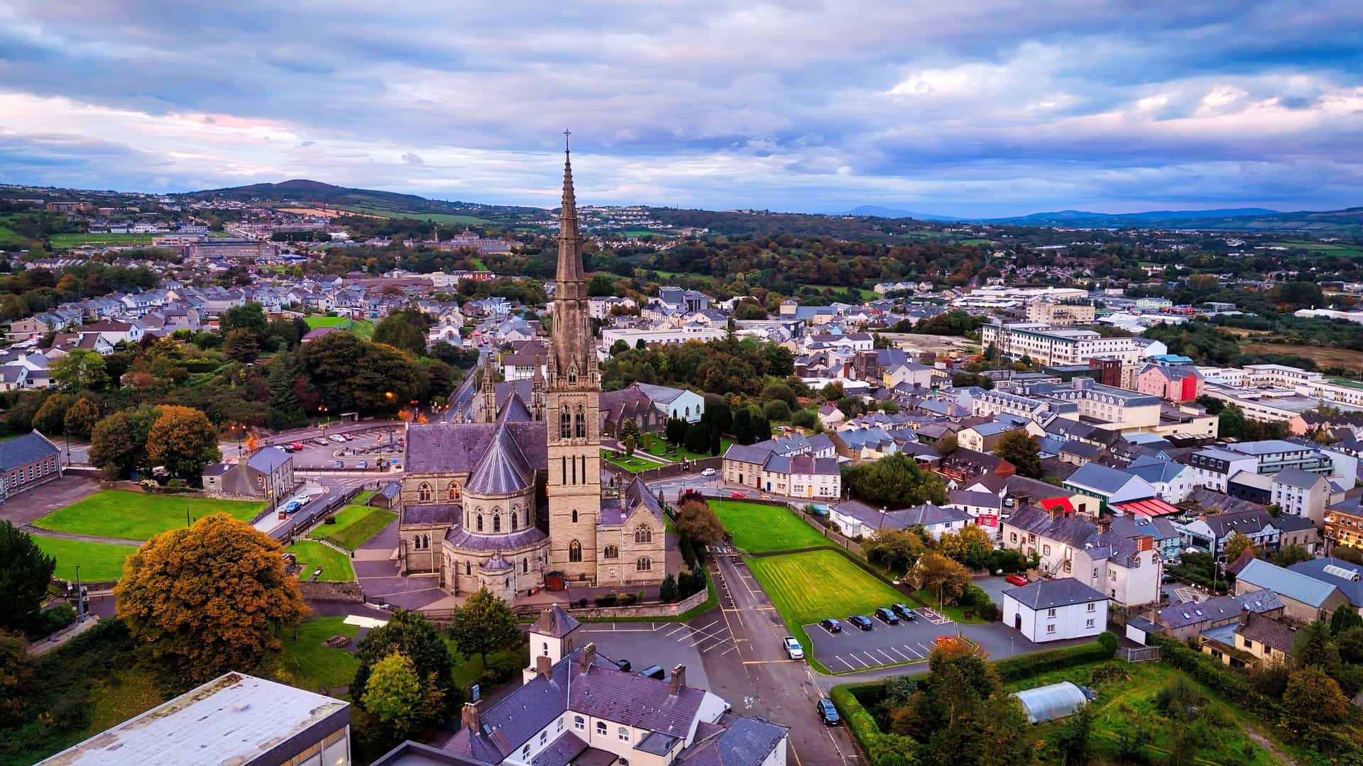 "An aerial view of Letterkenny, Ireland, showing the neo-gothic St. Eunan's Cathedral with its tall spire, surrounded by the town's buildings and green landscapes at sunset. "