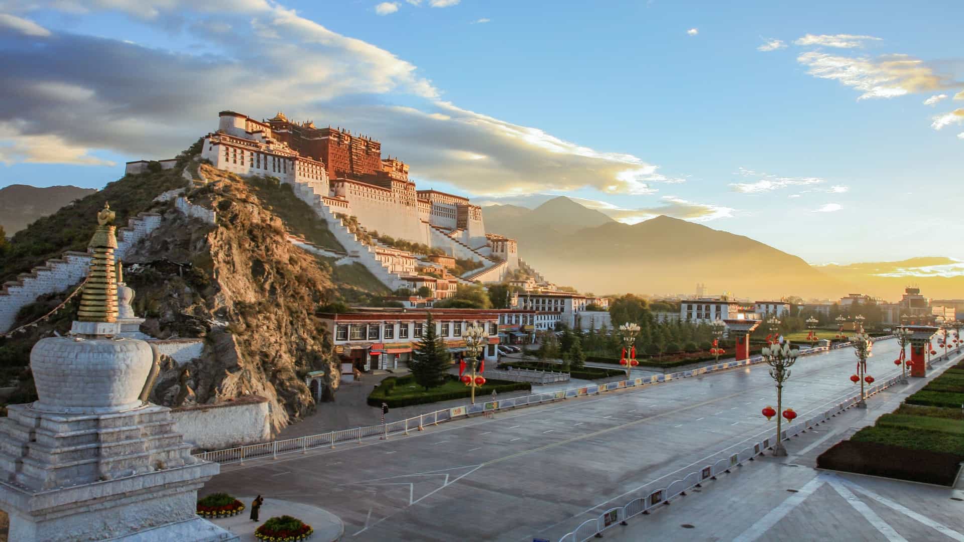 A beautiful sunset shot of the majestic Potala Palace in Lhasa, Tibet, a massive fortress built on a rocky hill with an empty road in the foreground and mountains in the distance.