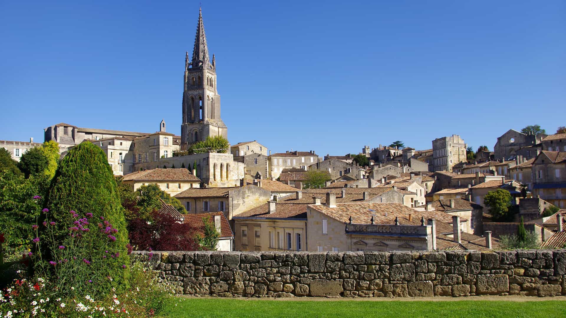 A picturesque view of the medieval town of Saint-Émilion near Libourne, with the Monolithic Church bell tower rising above the red rooftops against a clear blue sky.