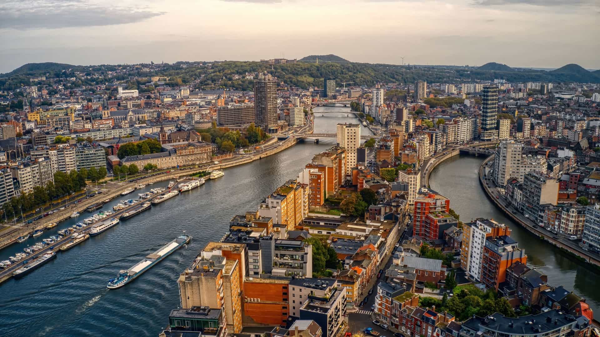 An aerial view of Liège, Belgium, showing the Meuse River winding through the cityscape with bridges, buildings, and barges under a hazy sky.