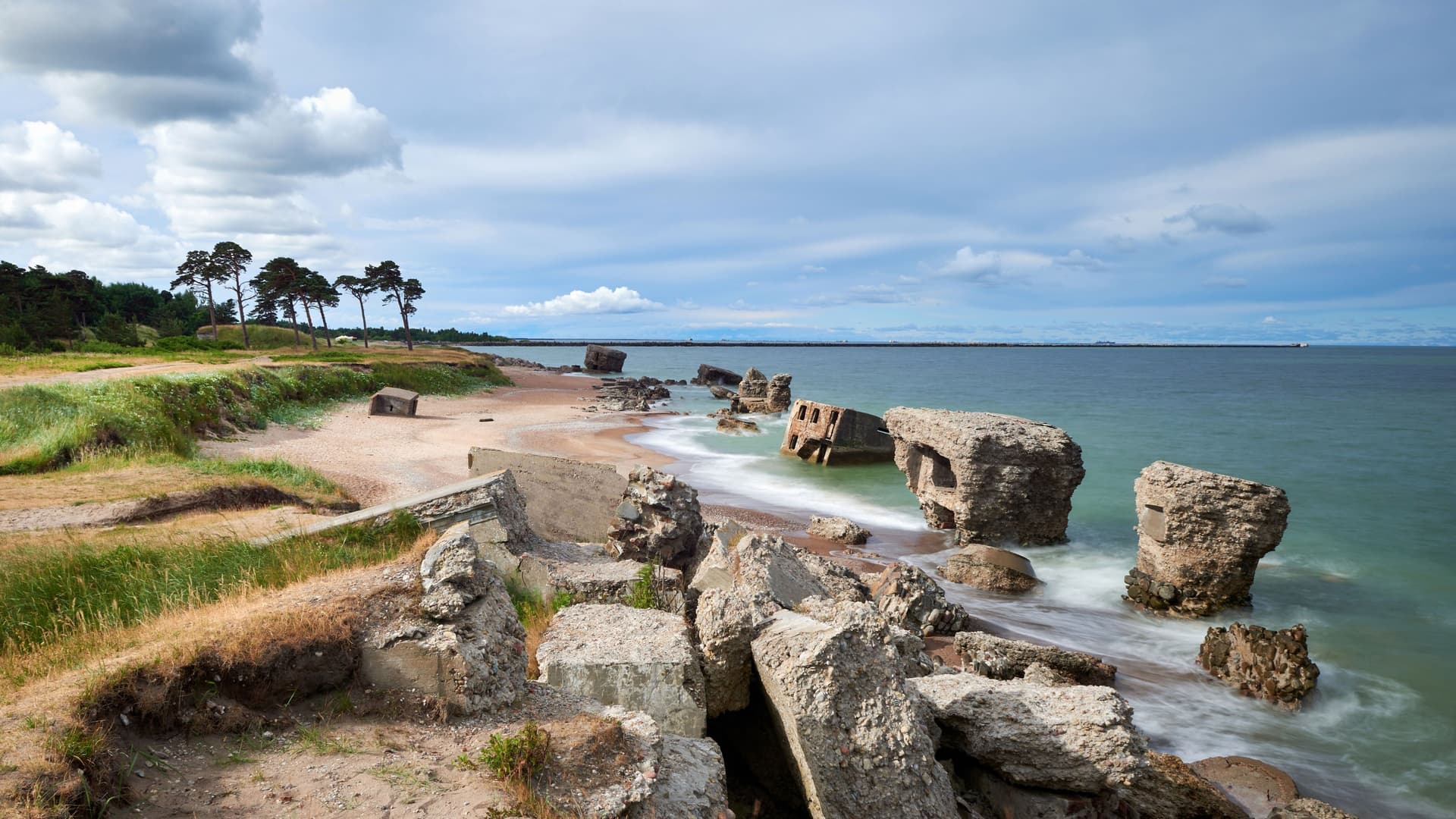 A panoramic shot of the scenic coast near Liepaja, Latvia, showing the ruins of concrete sea forts on the beach with waves crashing around them.