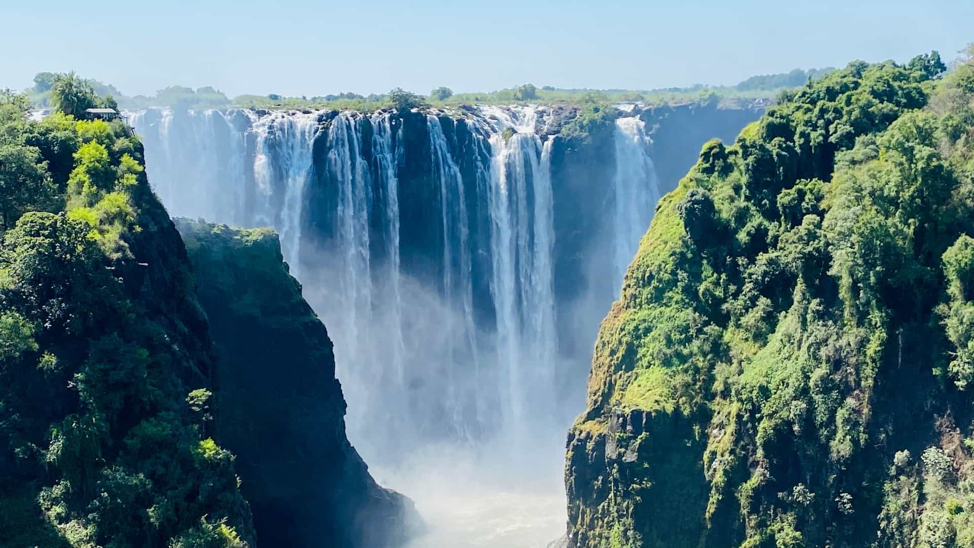 A huge curtain of Victoria Falls water plunges into a gorge surrounded by lush, mist-covered green cliffs near Livingstone, Zambia.