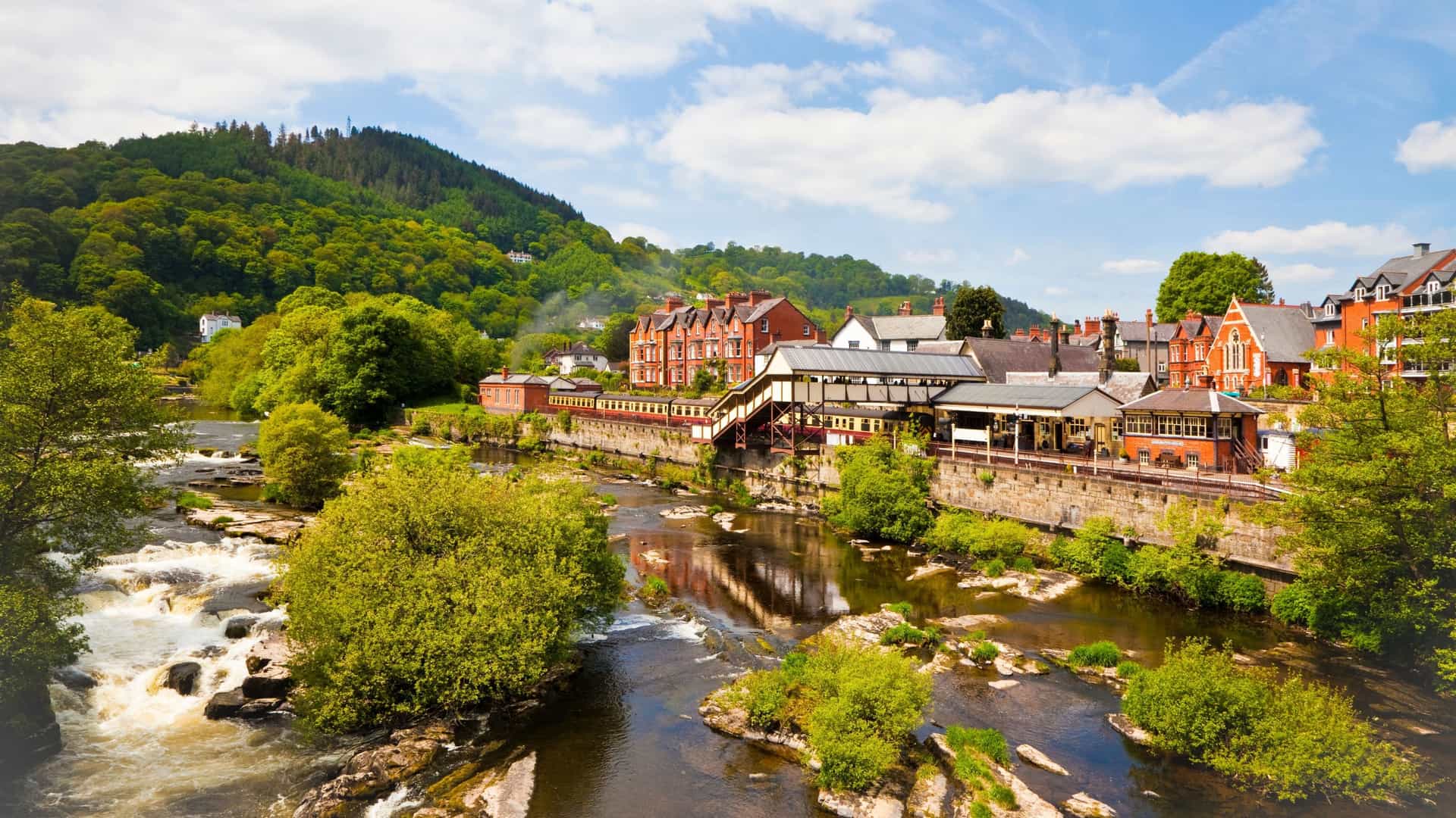 A beautiful daytime shot of the Llangollen railway station with its red-brick buildings, situated next to the rushing River Dee and surrounded by lush green hills in Wales.