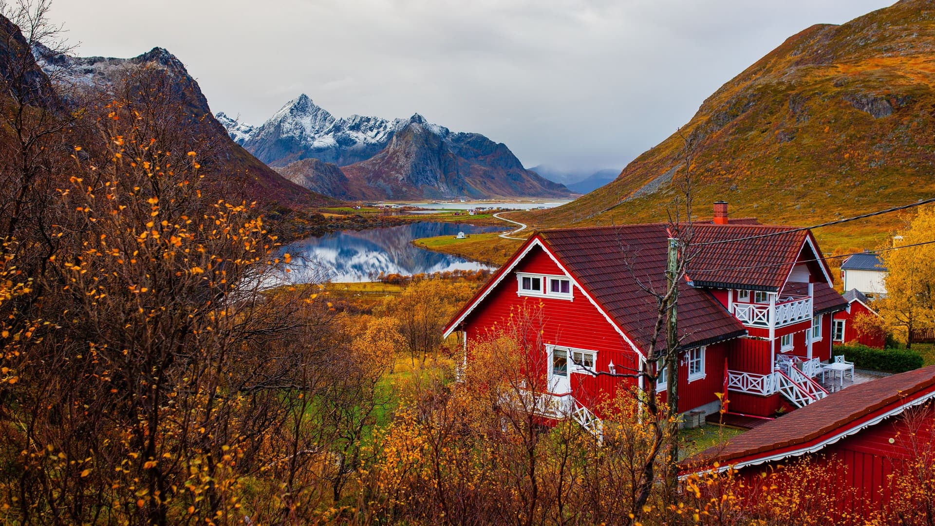 A breathtaking autumn view of a traditional red Norwegian house nestled among golden foliage near Loen, with majestic snow-capped mountains reflected in a calm lake.