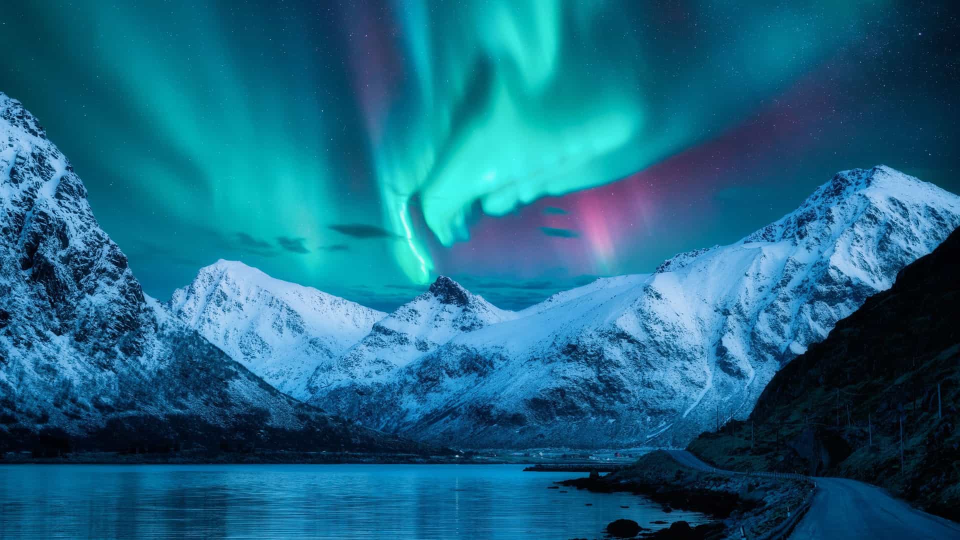 A dramatic night sky with a vibrant aurora borealis, or northern lights, over snow-covered mountains and a calm lake in the remote Lofoten Islands of Norway.