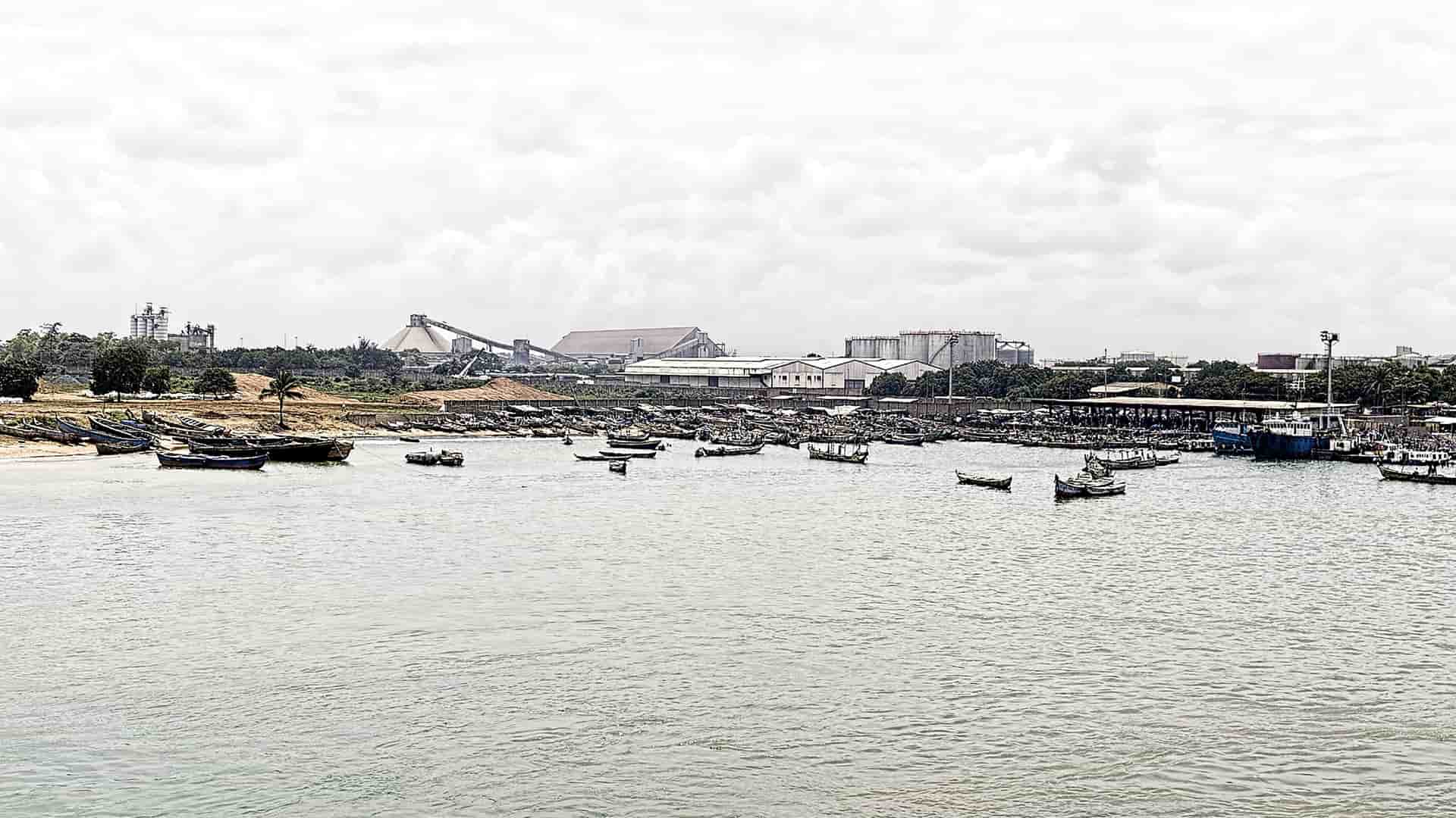 A busy and wide view of the Lomé seaport in Togo, West Africa, with numerous small fishing boats in the harbor and industrial buildings along the shoreline.