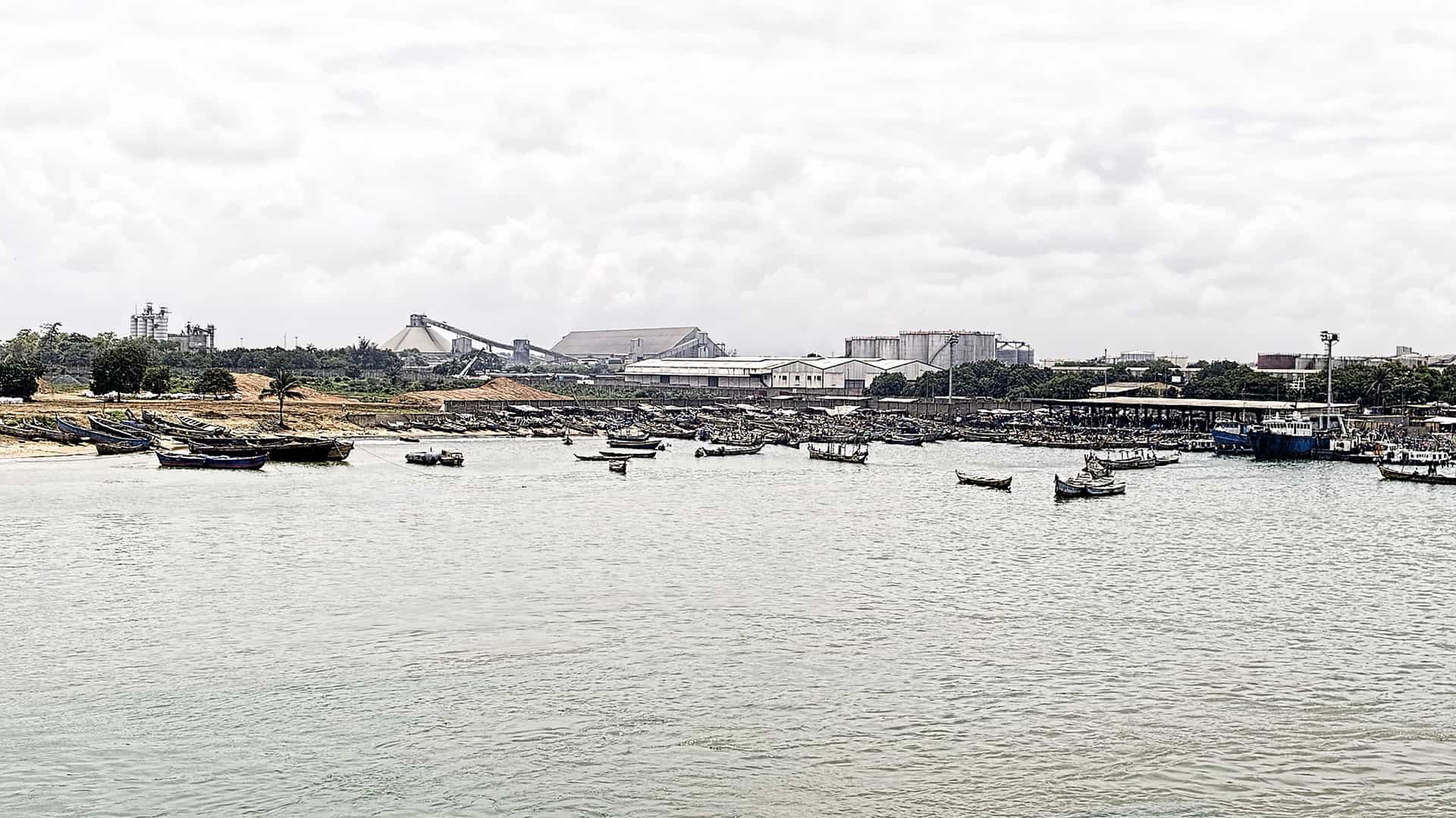 A busy and wide view of the Lomé seaport in Togo, West Africa, with numerous small fishing boats in the harbor and industrial buildings along the shoreline.