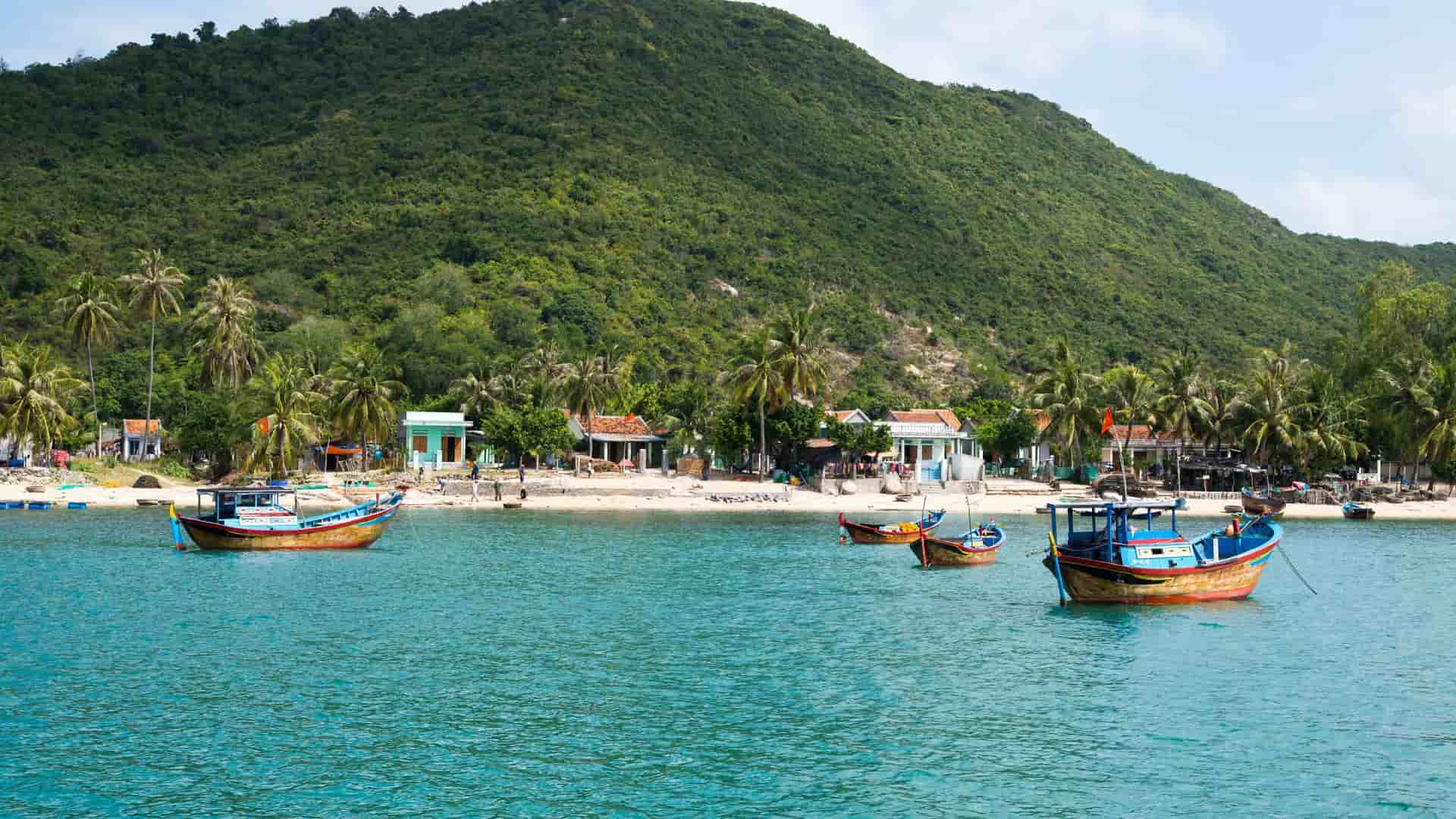 A serene tropical scene on Long Khanh Island in Vietnam, featuring traditional fishing boats floating in clear turquoise water in front of a small village and a lush green mountain.
