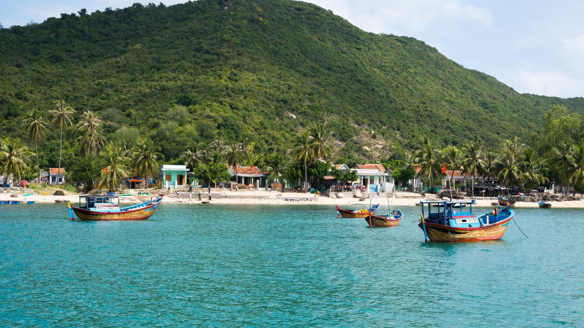 A serene tropical scene on Long Khanh Island in Vietnam, featuring traditional fishing boats floating in clear turquoise water in front of a small village and a lush green mountain.