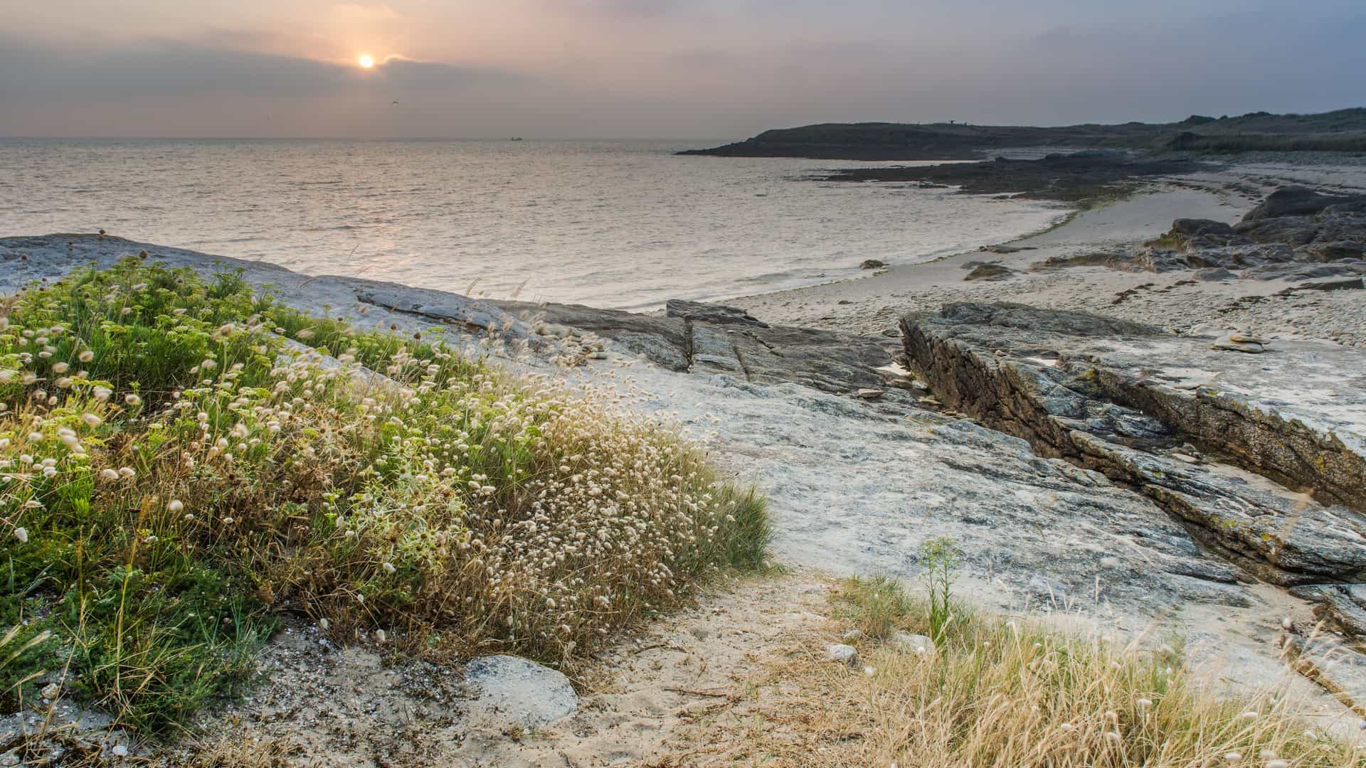 A serene sunset view over the rocky shoreline near Lorient, France, with a tranquil sea and sparse vegetation on a white-pebbled beach under a hazy sky.