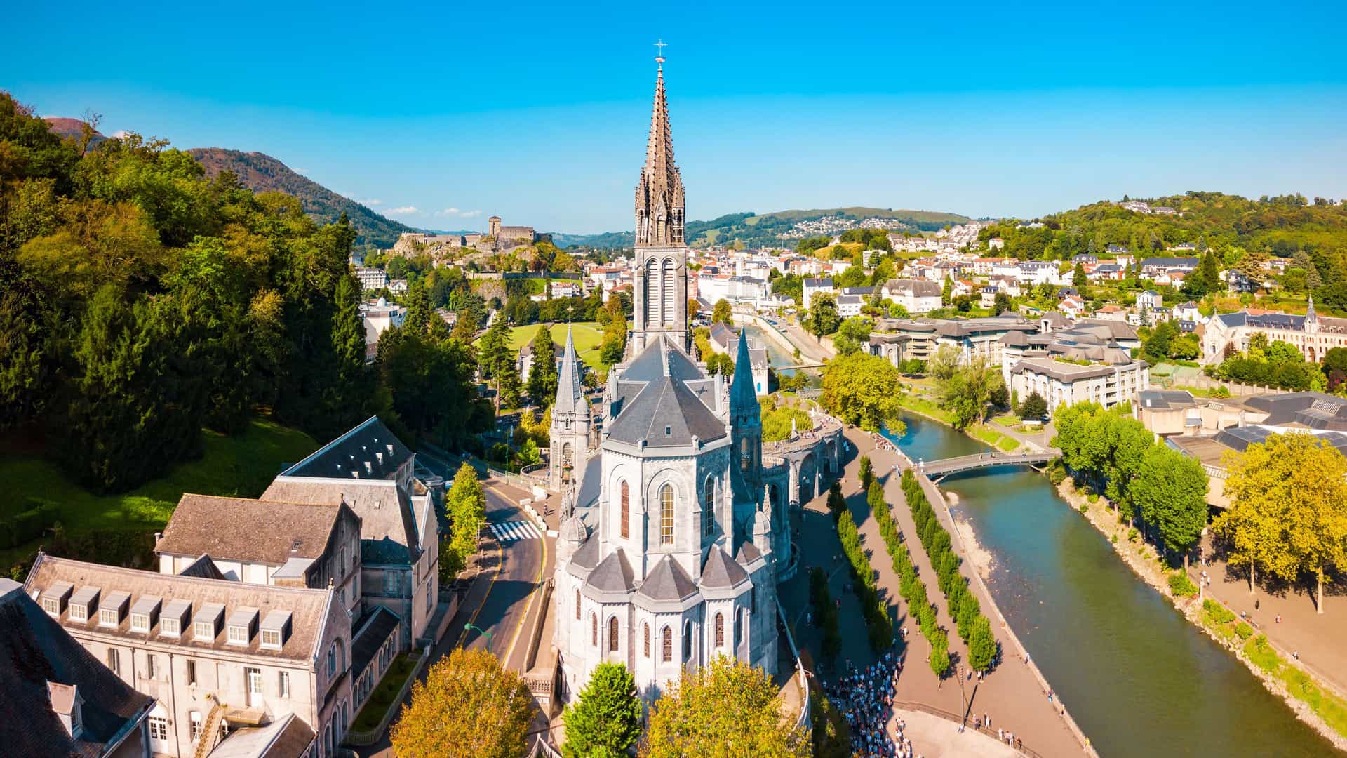"An aerial panoramic view of the Sanctuary of Our Lady of Lourdes in Lourdes, France, showing the famous basilica, the Gave de Pau river, and the surrounding Pyrenees mountains. "