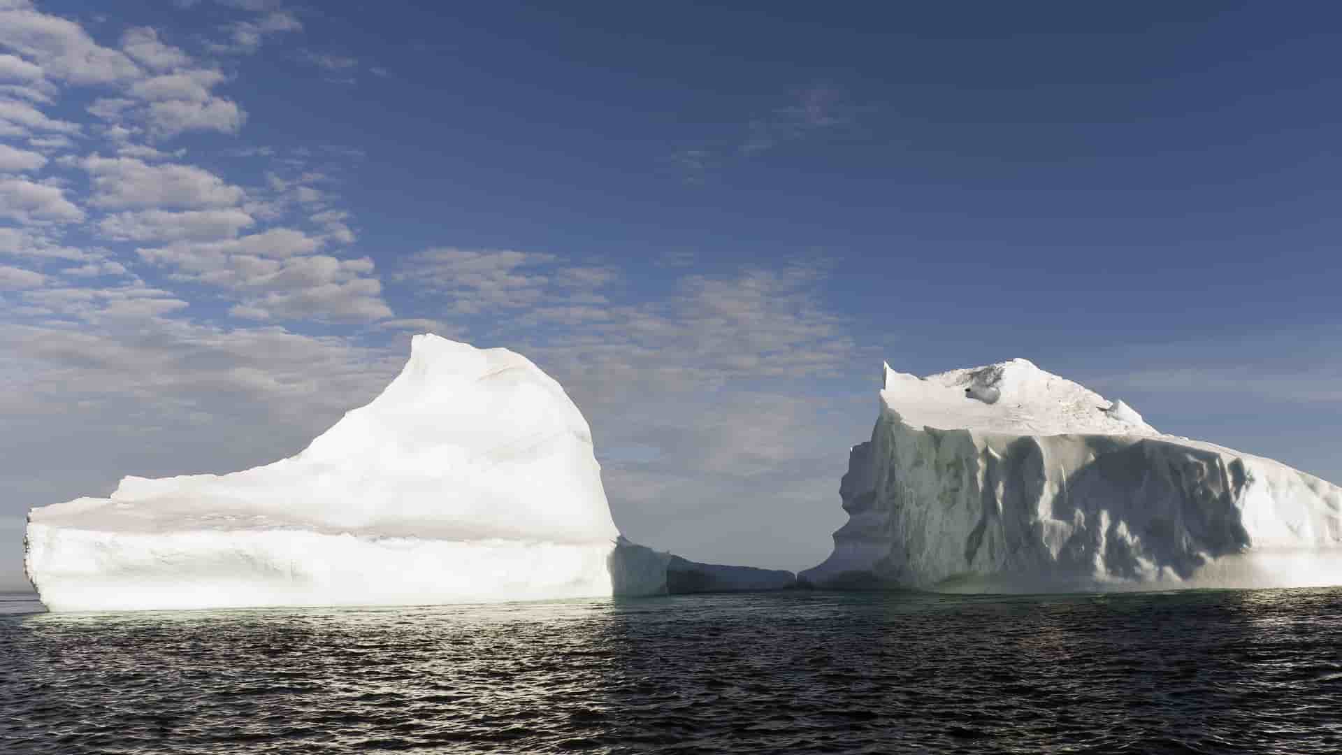 A breathtaking view of two massive, white icebergs floating in the dark ocean near the Lower Savage Islands of Nunavut, Canada, under a bright blue sky with scattered clouds.