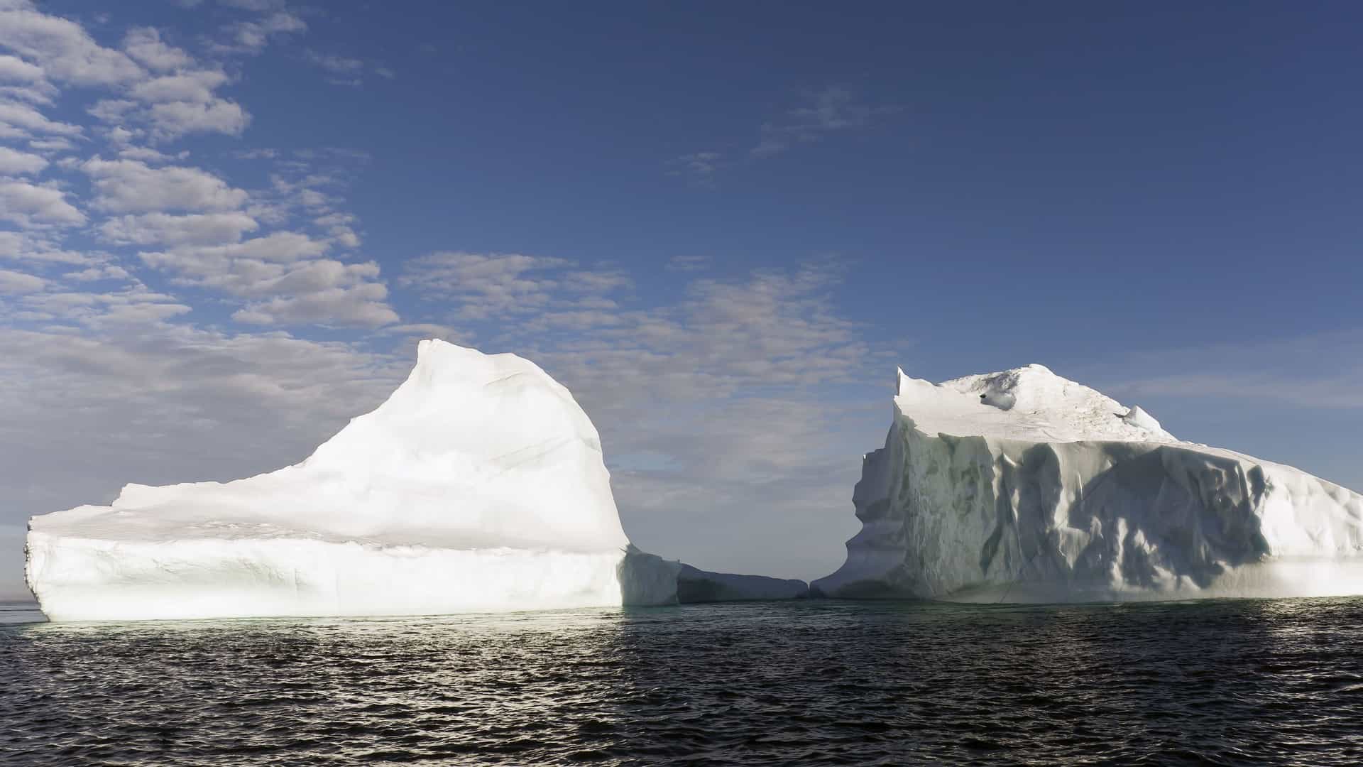 A breathtaking view of two massive, white icebergs floating in the dark ocean near the Lower Savage Islands of Nunavut, Canada, under a bright blue sky with scattered clouds.