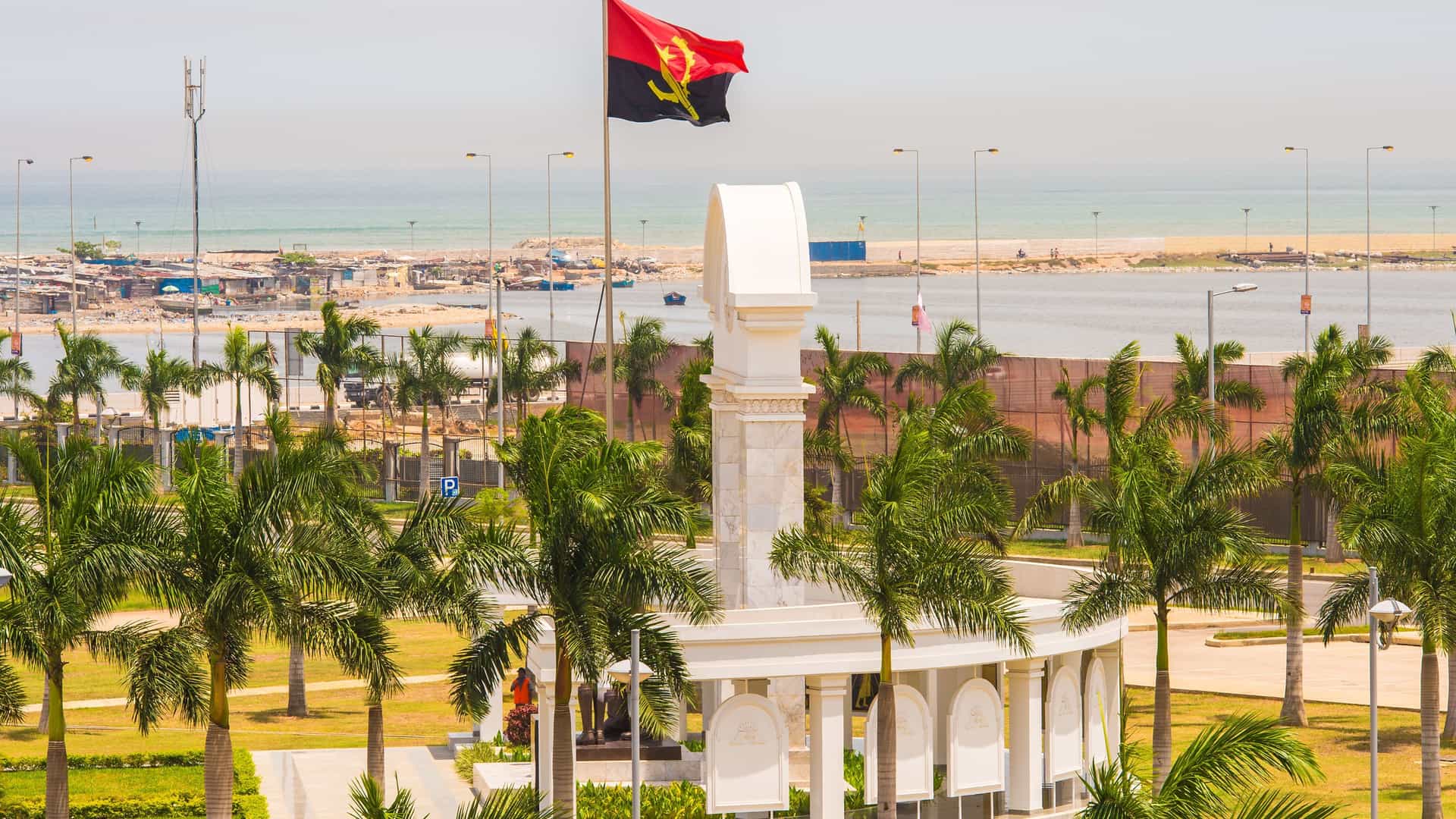 A scenic view of the waterfront in Luanda, Angola, with a prominent monument, palm trees, and the national flag flying high against the backdrop of the Atlantic Ocean.