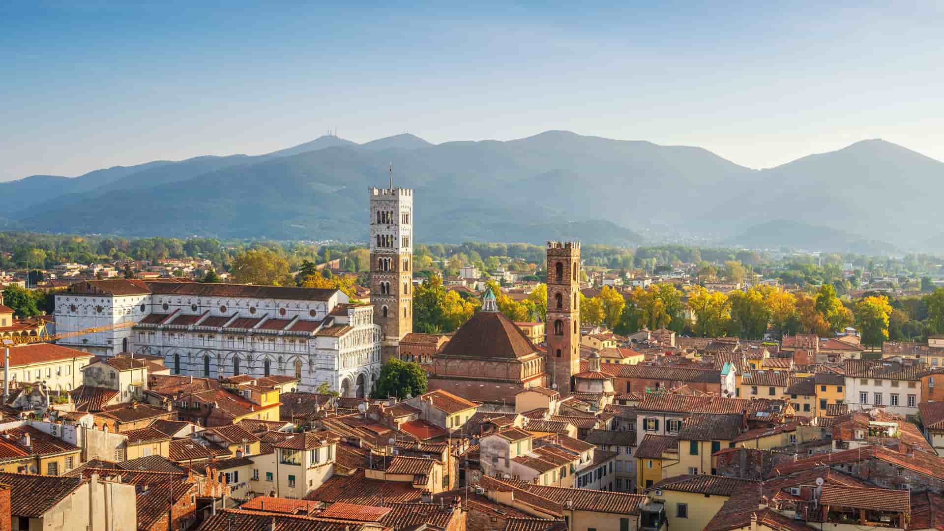 "An aerial view of the historic city of Lucca, Italy, with its terracotta rooftops, prominent bell towers, and a backdrop of rolling Tuscan hills. "