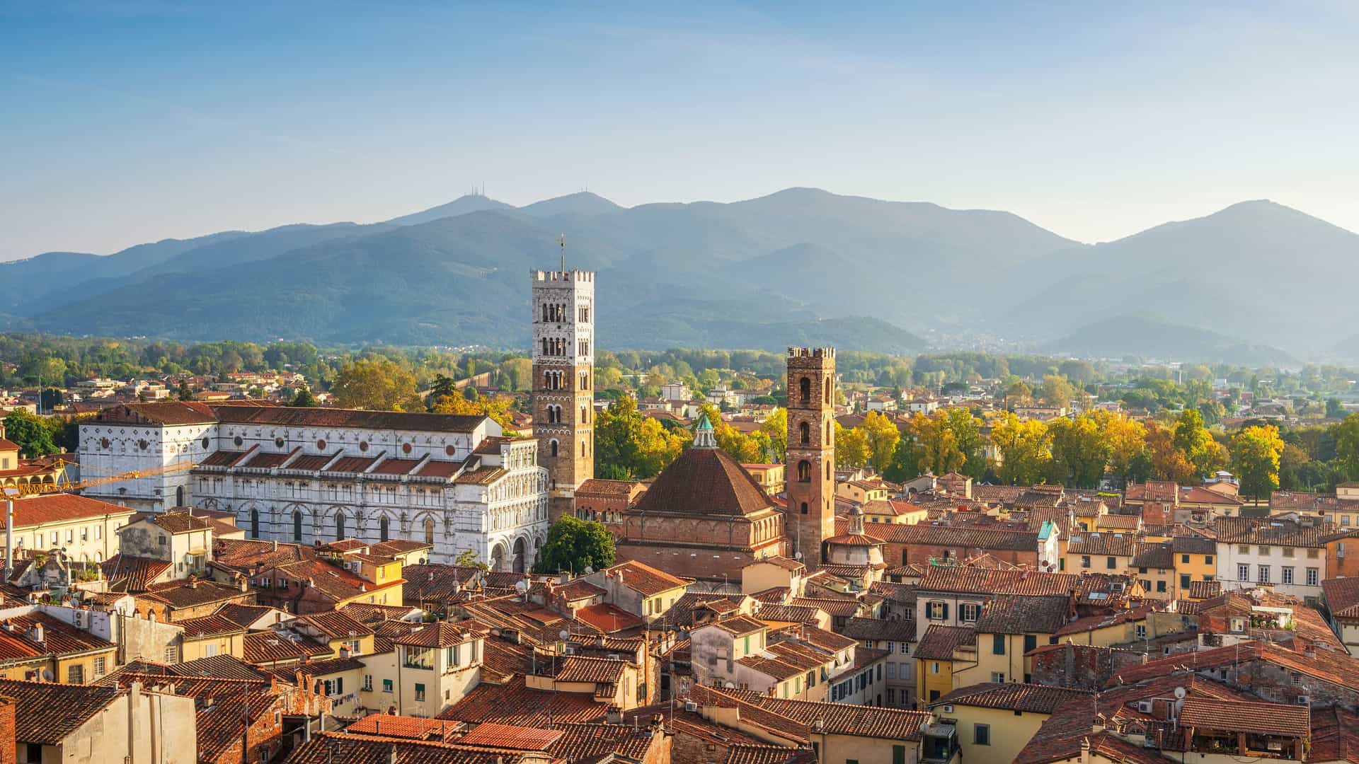 "An aerial view of the historic city of Lucca, Italy, with its terracotta rooftops, prominent bell towers, and a backdrop of rolling Tuscan hills. "
