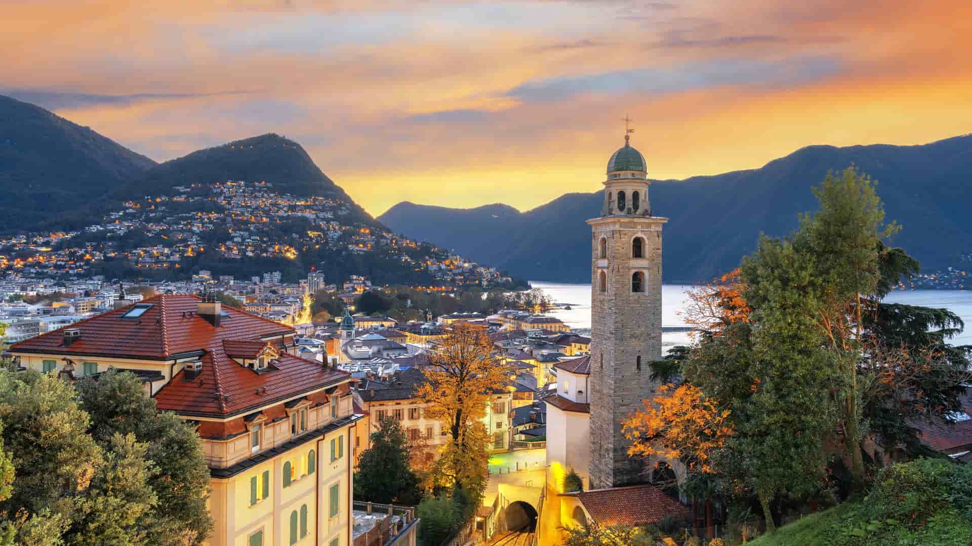 "An elevated sunset view of Lugano, Switzerland, highlighting the beautiful bell tower of the Church of Santa Maria degli Angeli, with Lake Lugano and the surrounding mountains in the background. "
