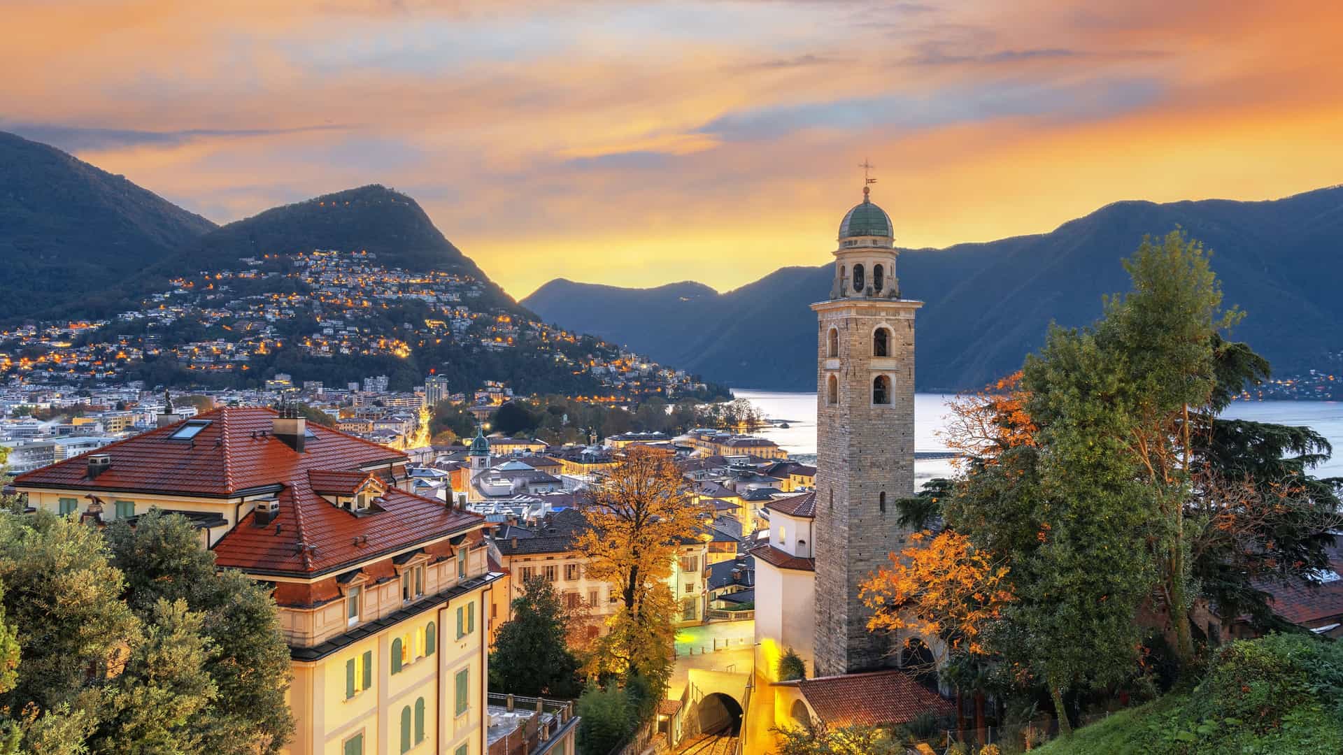 "An elevated sunset view of Lugano, Switzerland, highlighting the beautiful bell tower of the Church of Santa Maria degli Angeli, with Lake Lugano and the surrounding mountains in the background. "