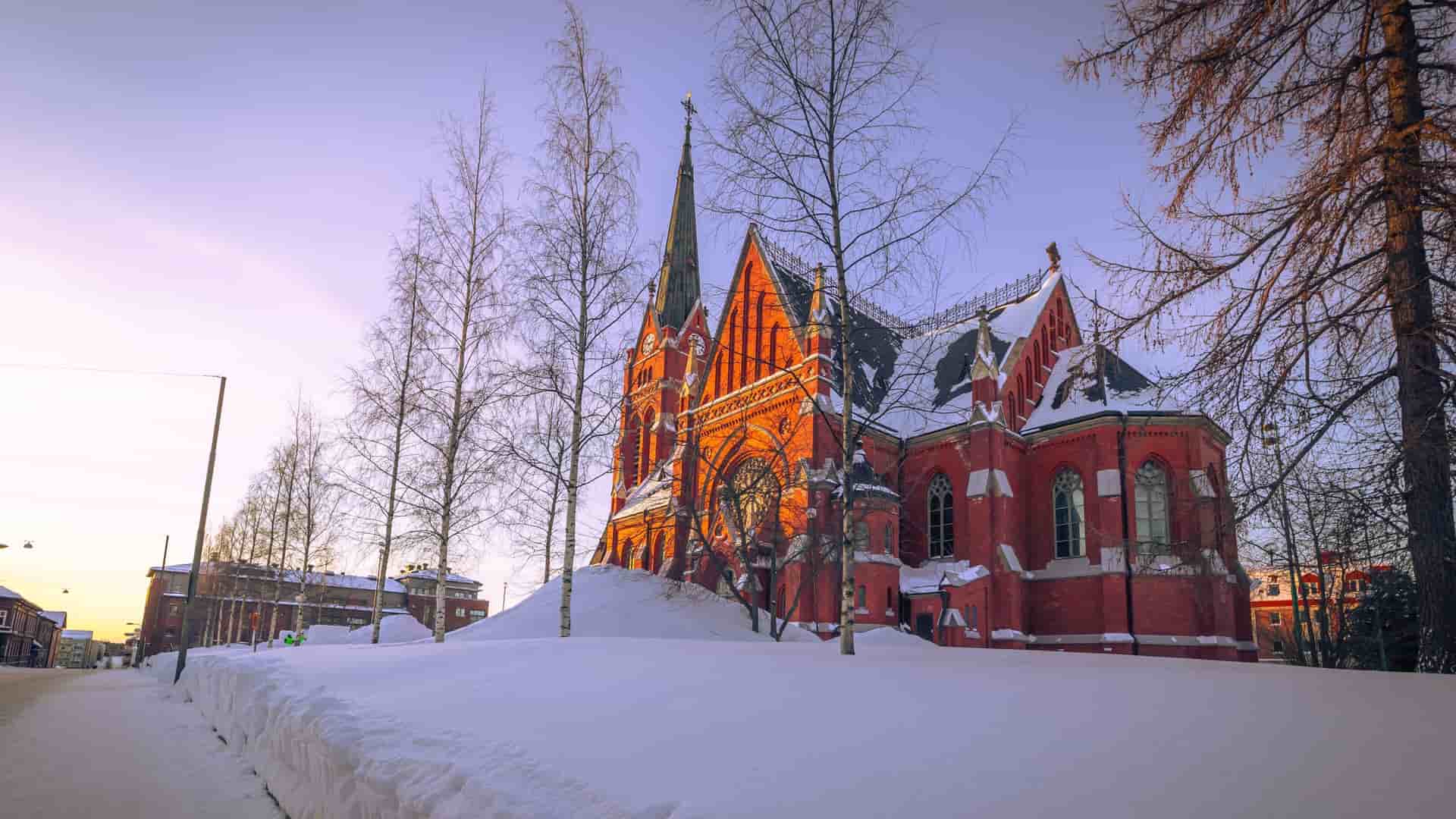 A winter image of the red-brick Luleå Cathedral in Sweden, covered in deep snow and surrounded by bare trees, glowing in the warm light of the setting sun.