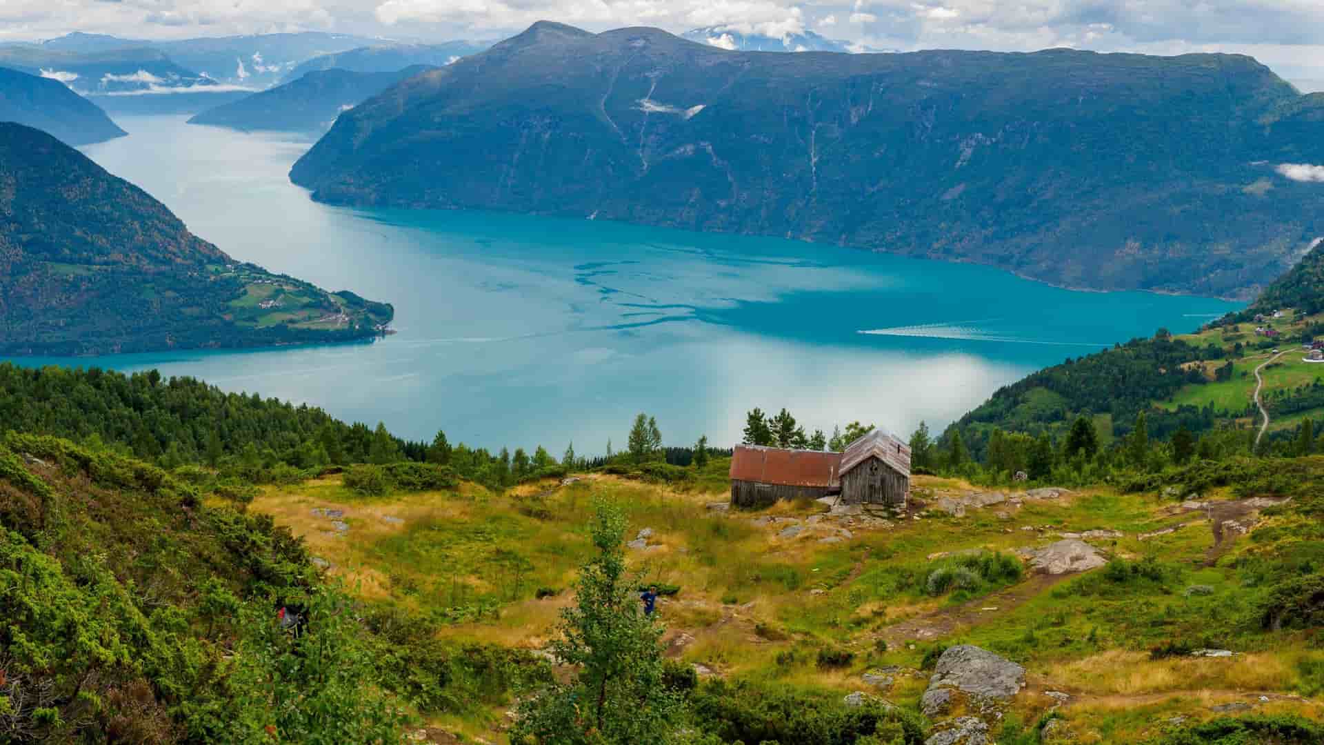 A breathtaking panoramic shot of the Lustrafjord in Norway, showcasing the turquoise waters, lush green landscape with a rustic hut, and majestic mountains under a cloudy sky.