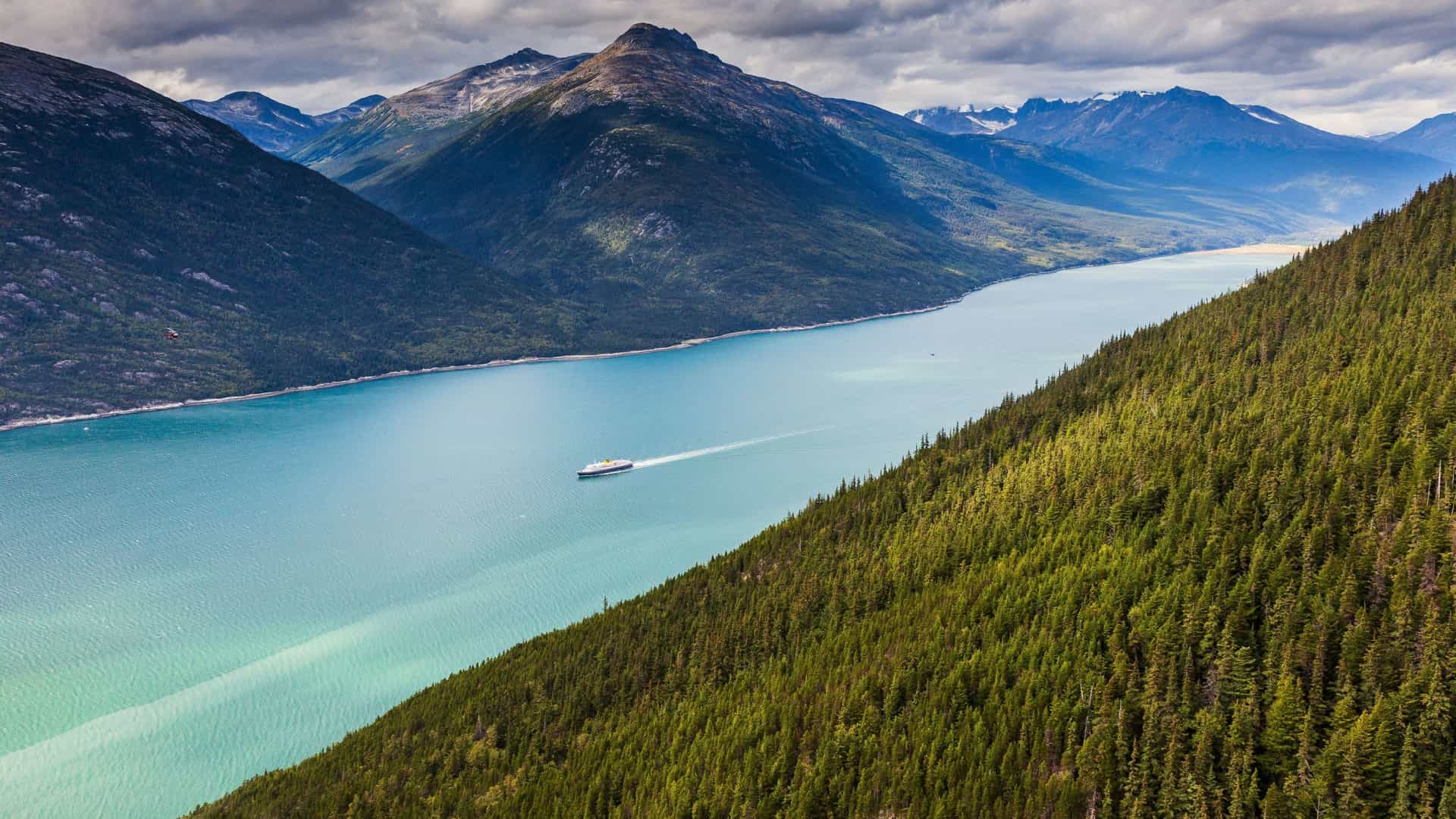 A high-angle view of a cruise ship sailing through the glacial Lynn Canal, a stunning fjord in Alaska, surrounded by majestic mountains and lush green forests.