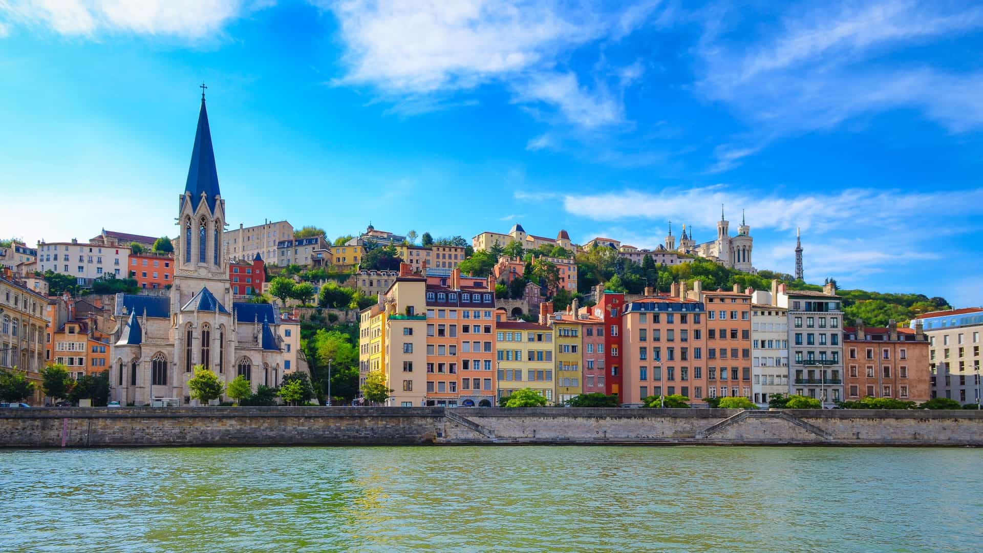 A panoramic shot of the historic cityscape of Lyon, France, featuring colorful buildings along the Saône River and the Notre-Dame de Fourvière Basilica perched on the hilltop.