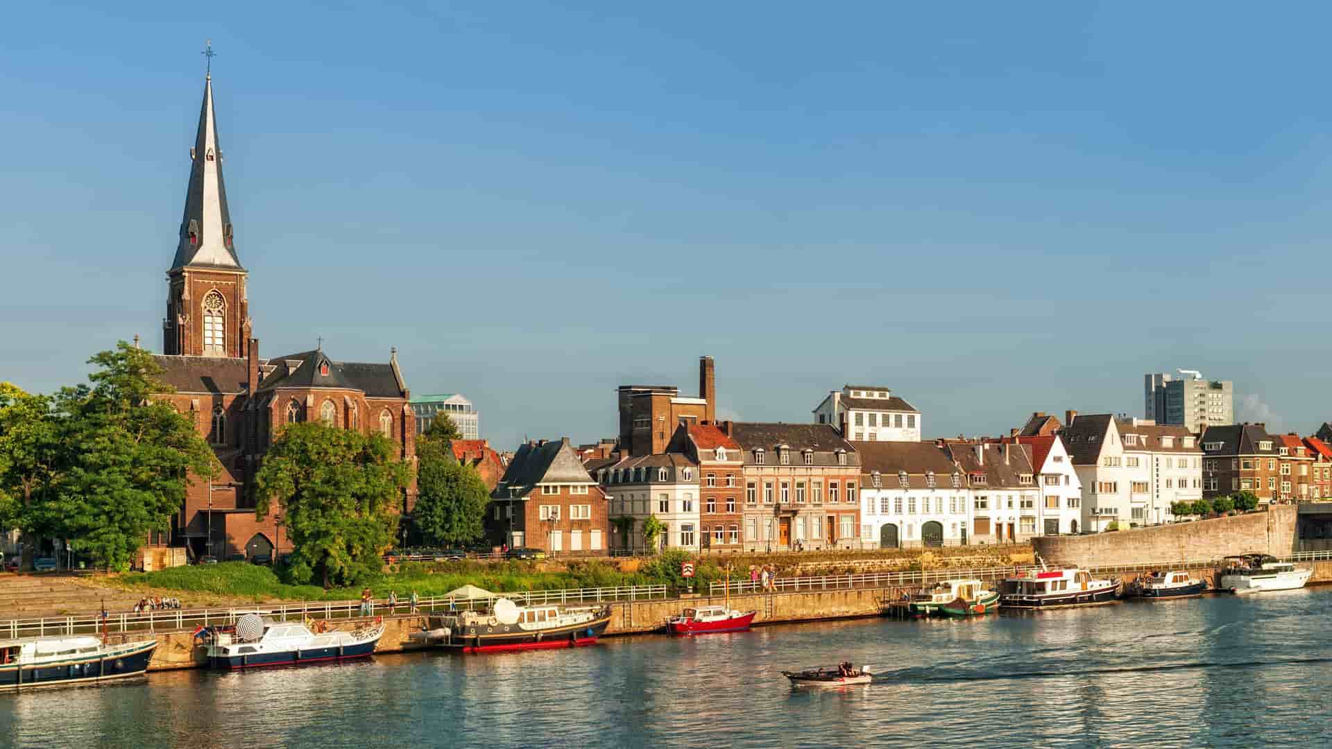 A scenic view of Maastricht, Netherlands, with the historic St. Servatius Church and traditional buildings lining the Maas River, with boats docked along the waterfront.