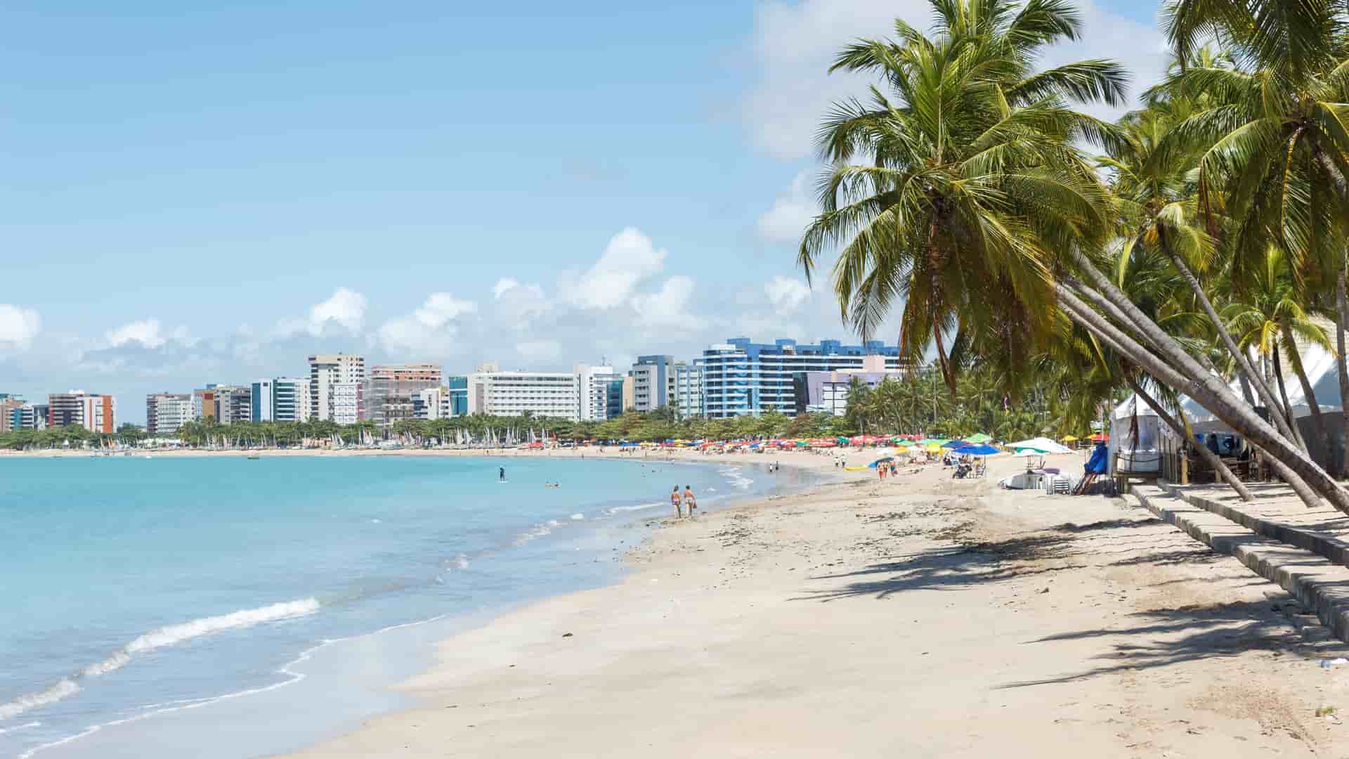 A scenic shot of the beautiful Maceió beach in Brazil, with a calm turquoise sea, a stretch of golden sand, and tall palm trees lining the shore with the city skyline in the distance.