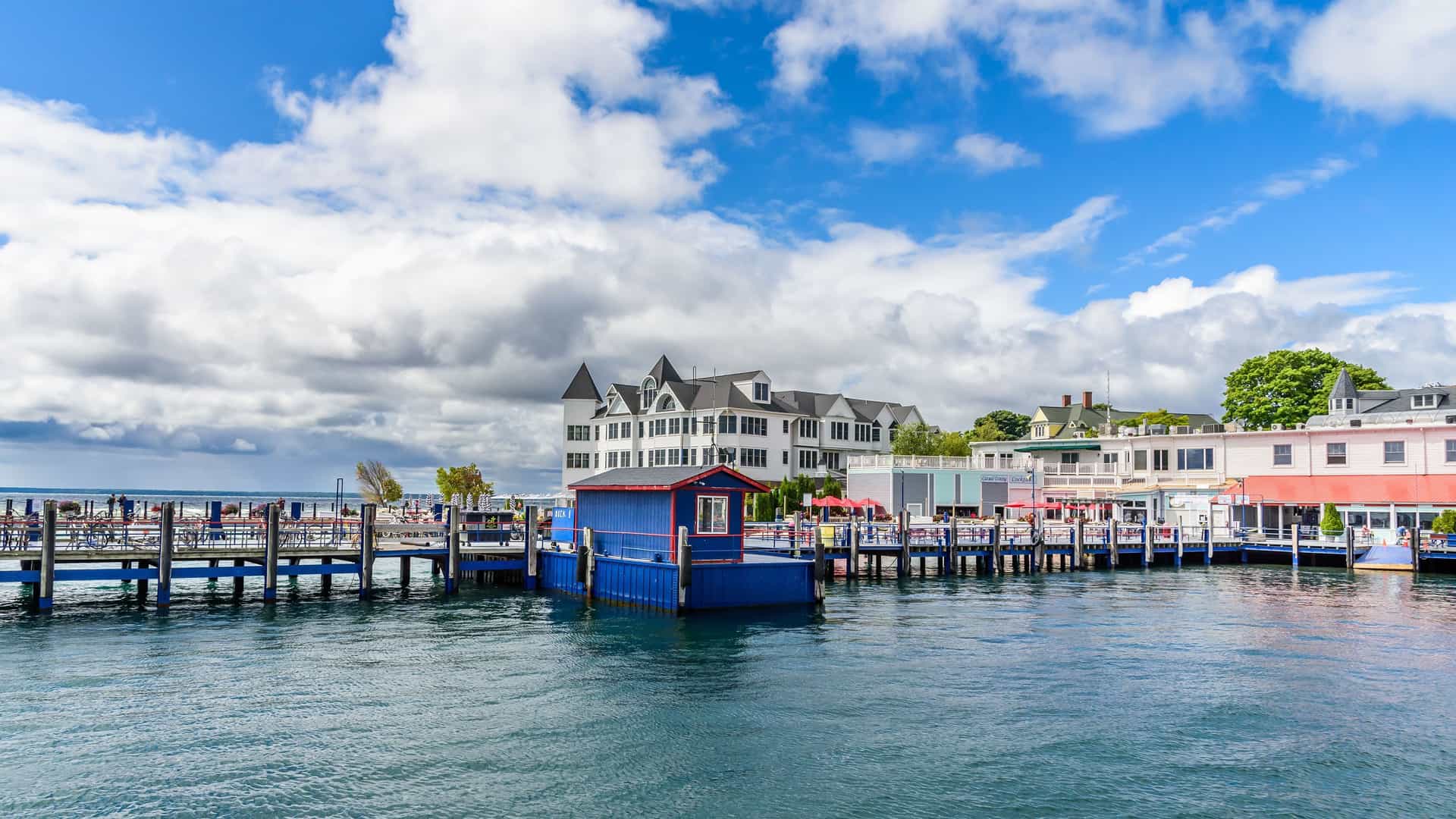 A vibrant shot of the waterfront on Mackinac Island, Michigan, with a colorful blue boathouse and a row of classic white and red buildings under a bright blue sky with puffy white clouds.