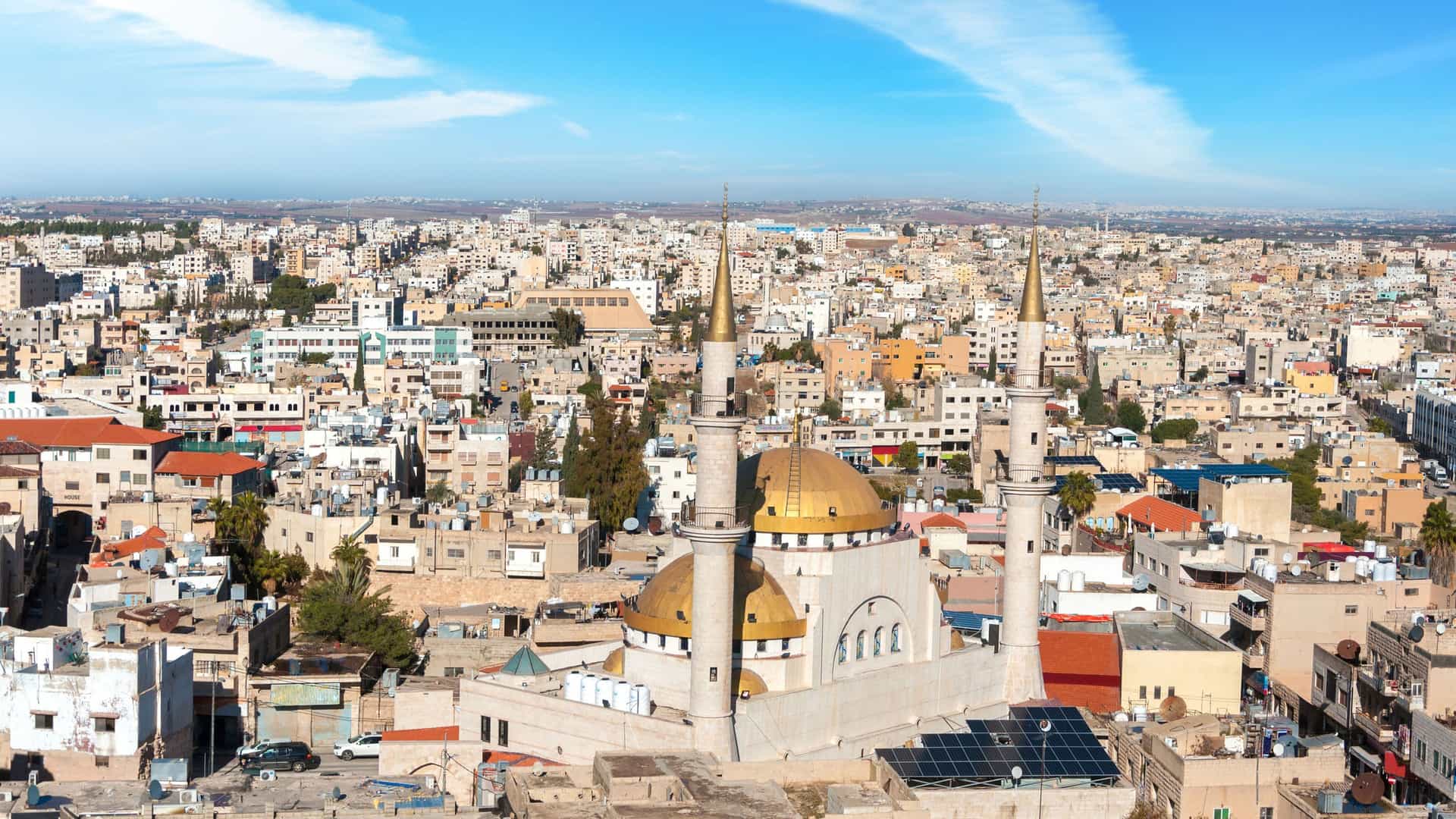 A high-angle view of the bustling city of Madaba, Jordan, with a beautiful mosque featuring golden domes and minarets rising above the surrounding traditional buildings.