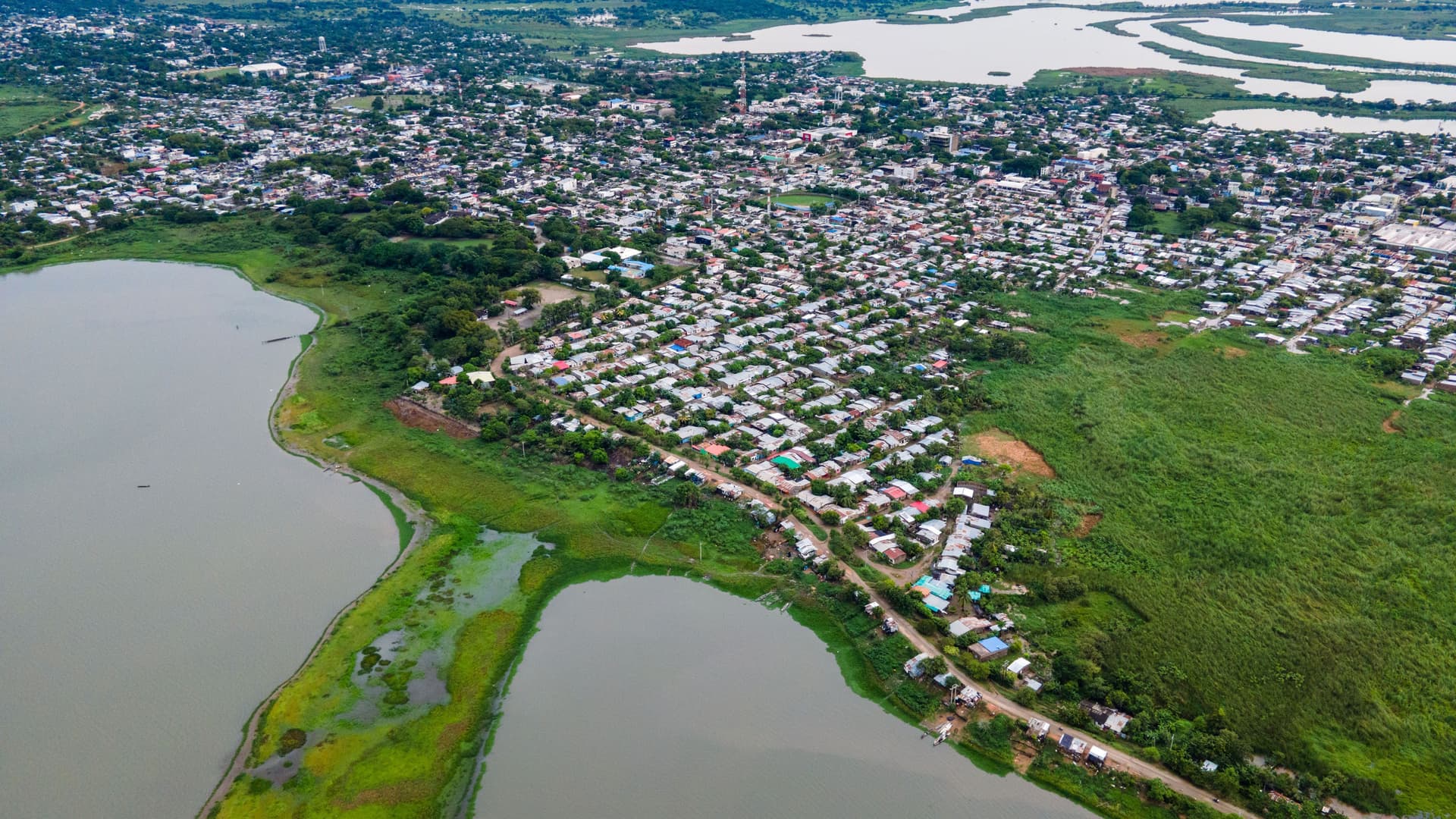 Aerial view of Magangué, Colombia, a densely built town with colorful roofs, bordered by a murky river/lake and lush green land, under an overcast sky.
