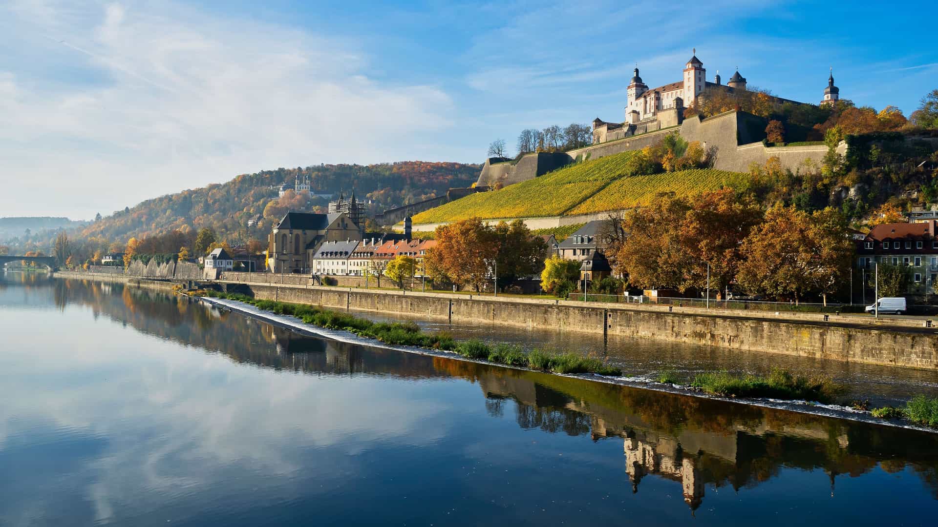 A panoramic shot of the historic Marienberg Fortress overlooking the Main River, with the scenic Würzburg cityscape reflecting in the water during autumn.
