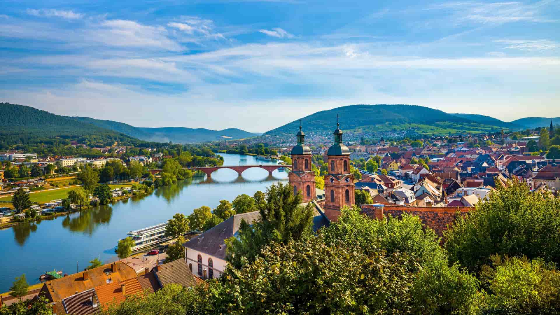 A beautiful village view on the Main River, with historic churches, a red bridge, and lush green hills under a bright blue sky.