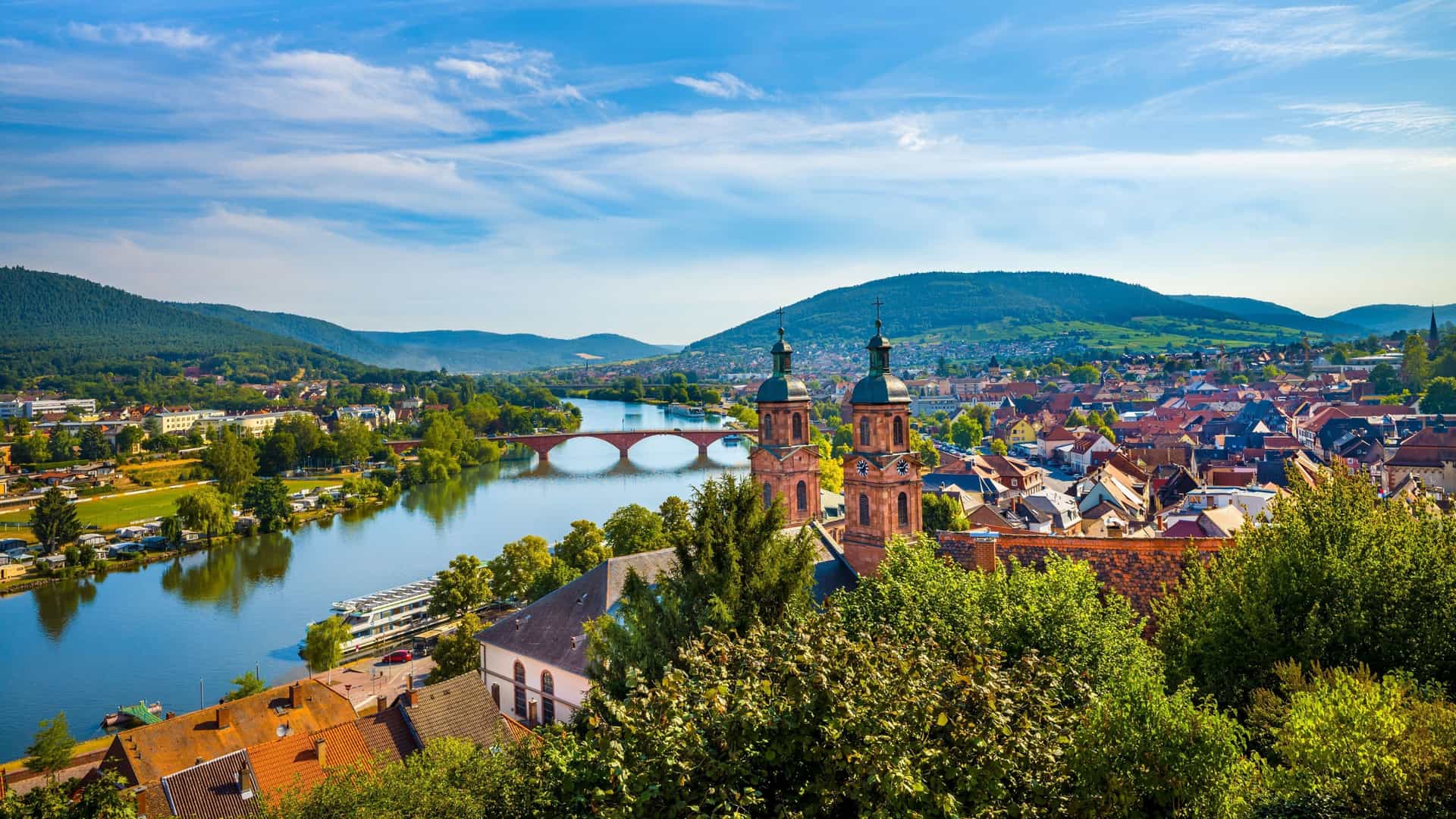 A beautiful village view on the Main River, with historic churches, a red bridge, and lush green hills under a bright blue sky.