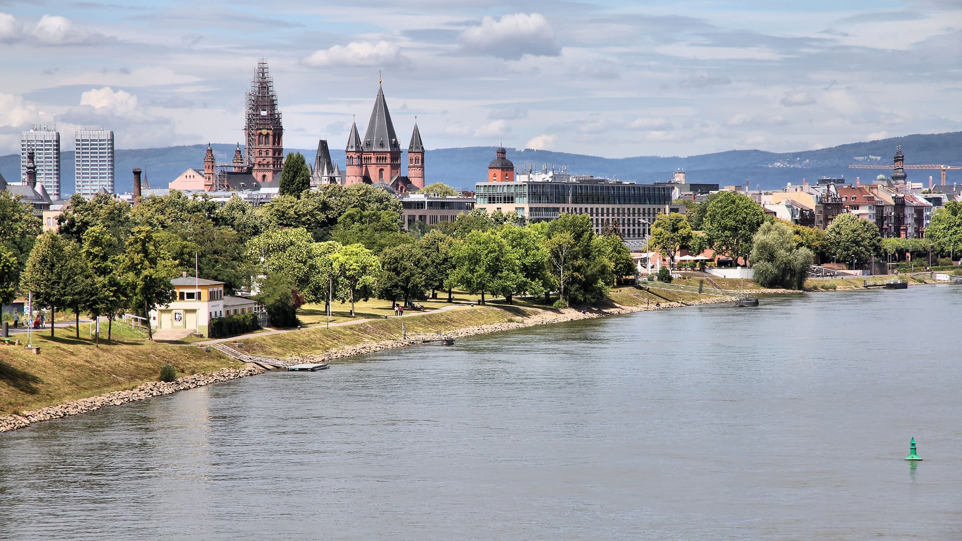 A cityscape of Mainz, Germany, from across the Rhine River, showcasing historic buildings, the distinctive cathedral, and green parkland along the waterfront.