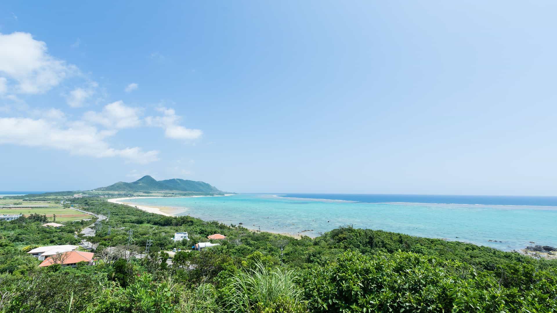 A stunning aerial shot of the coastline in Maizuru, Japan, with turquoise water, a sandy beach, and a lush green landscape under a clear blue sky.