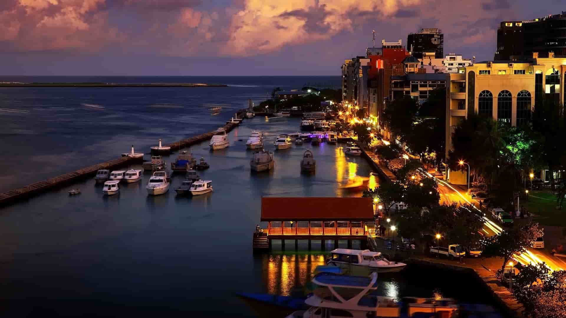 A nighttime cityscape view of Malé, Maldives, with boats in the harbor, city lights reflecting on the water, and a vibrant street scene.