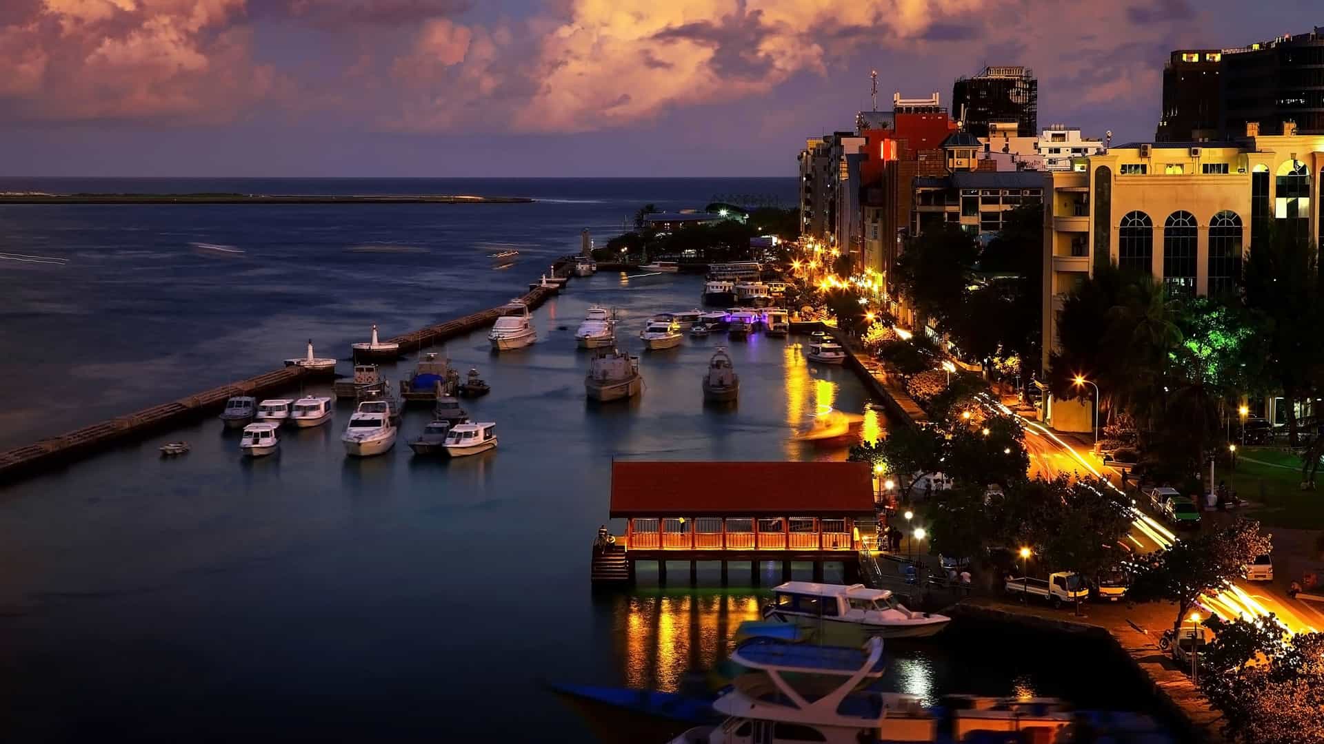 A nighttime cityscape view of Malé, Maldives, with boats in the harbor, city lights reflecting on the water, and a vibrant street scene.
