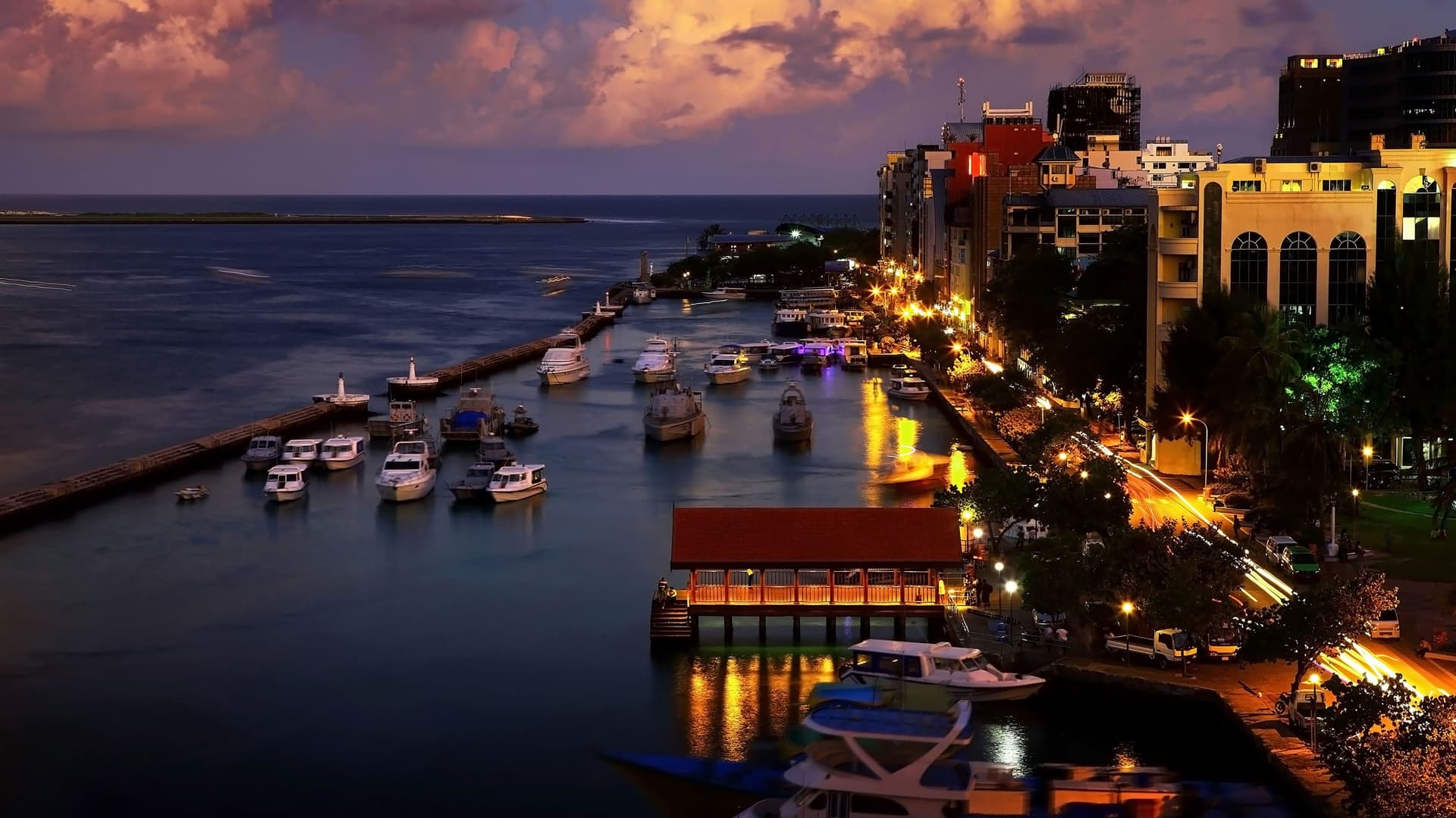 A nighttime cityscape view of Malé, Maldives, with boats in the harbor, city lights reflecting on the water, and a vibrant street scene.