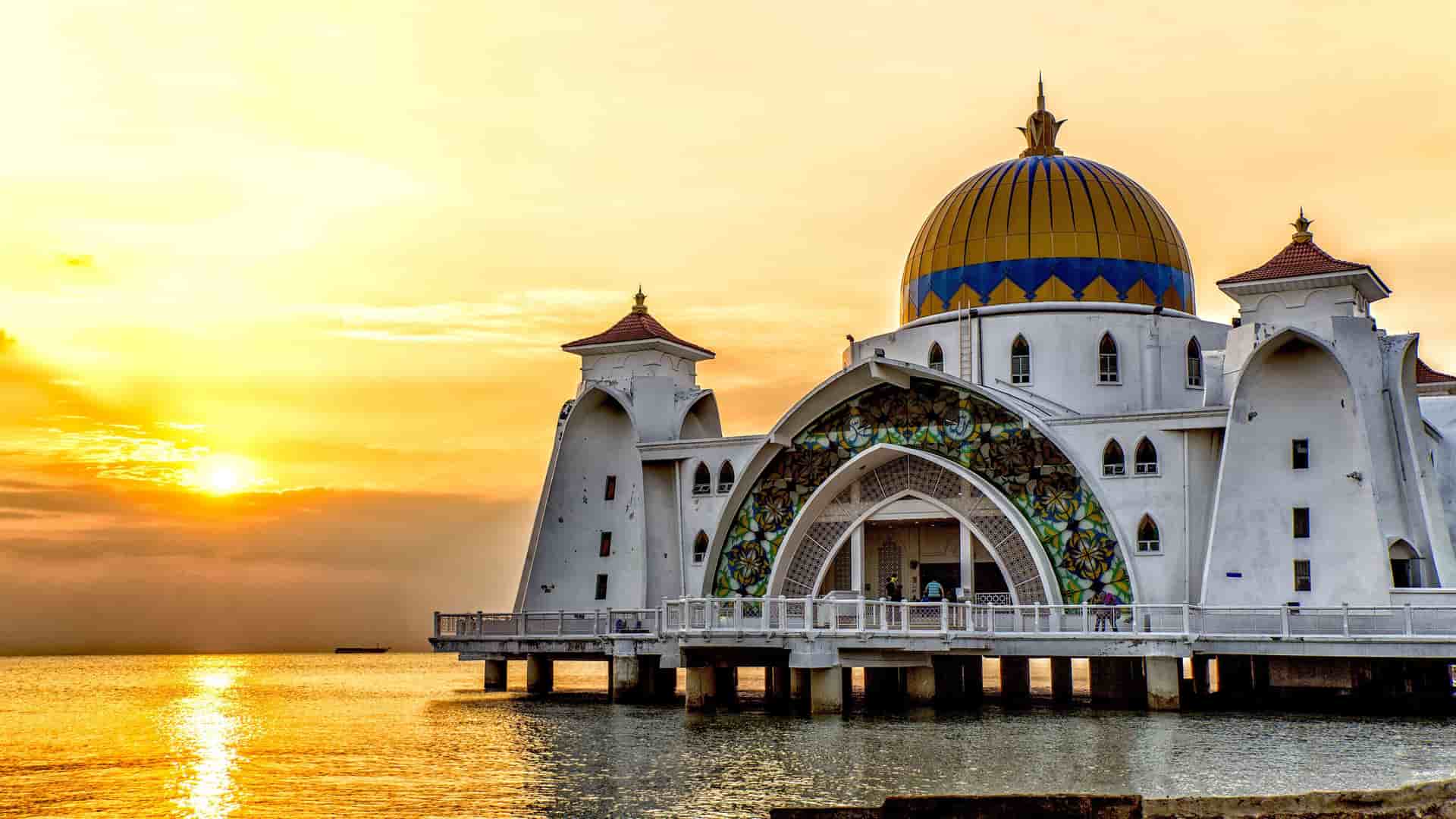 A dramatic sunset shot of the Malacca Straits Mosque in Malacca, Malaysia, with its dome and minarets reflecting on the golden water.
