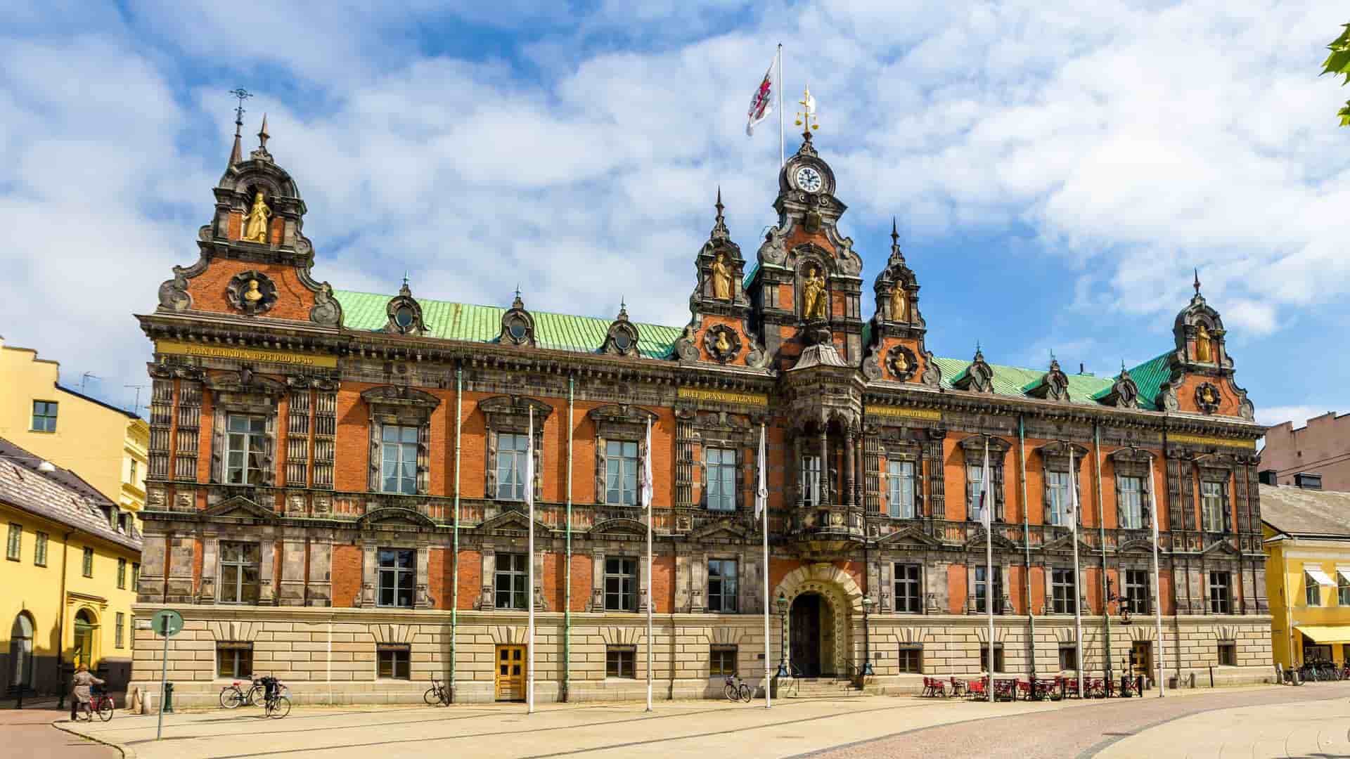 A frontal shot of the historic Malmö City Hall, a grand building with an ornate brick facade, a clock tower, and statues under a bright blue sky.