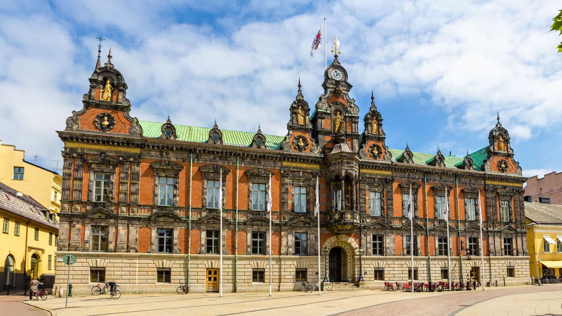 A frontal shot of the historic Malmö City Hall, a grand building with an ornate brick facade, a clock tower, and statues under a bright blue sky.