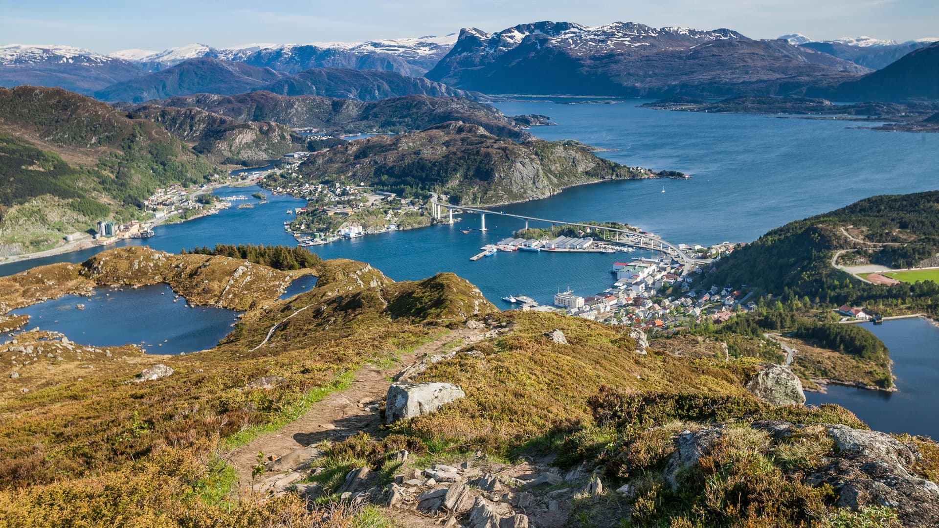A breathtaking panoramic shot of Måløy, Norway, from a mountain trail, with the town, its bridge, and the surrounding fjord and snow-capped mountains.