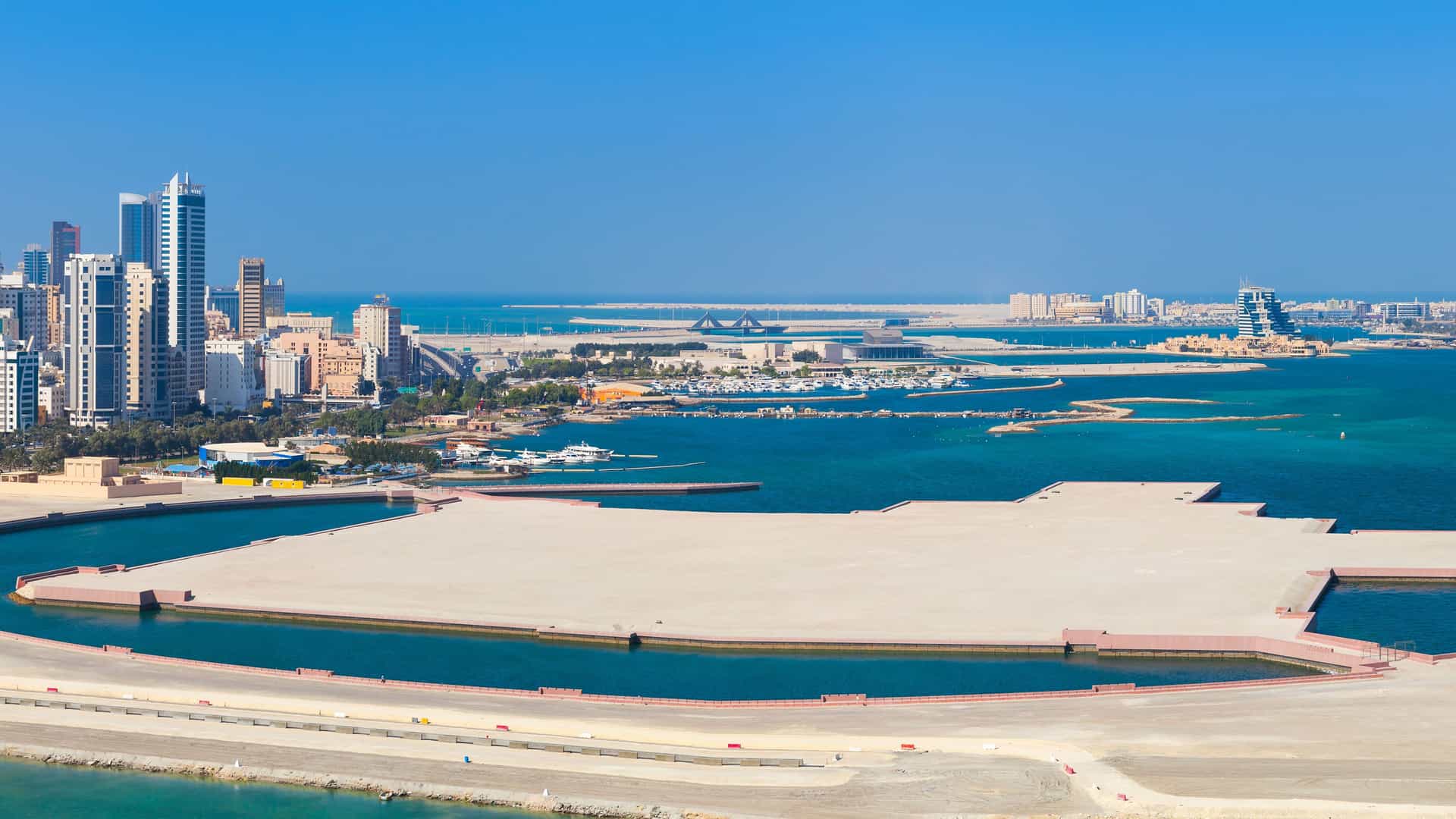 A clear, sunny aerial shot of the city of Manama, Bahrain, with its modern skyline, a large bay with multiple docks, and a pristine blue ocean in the background.