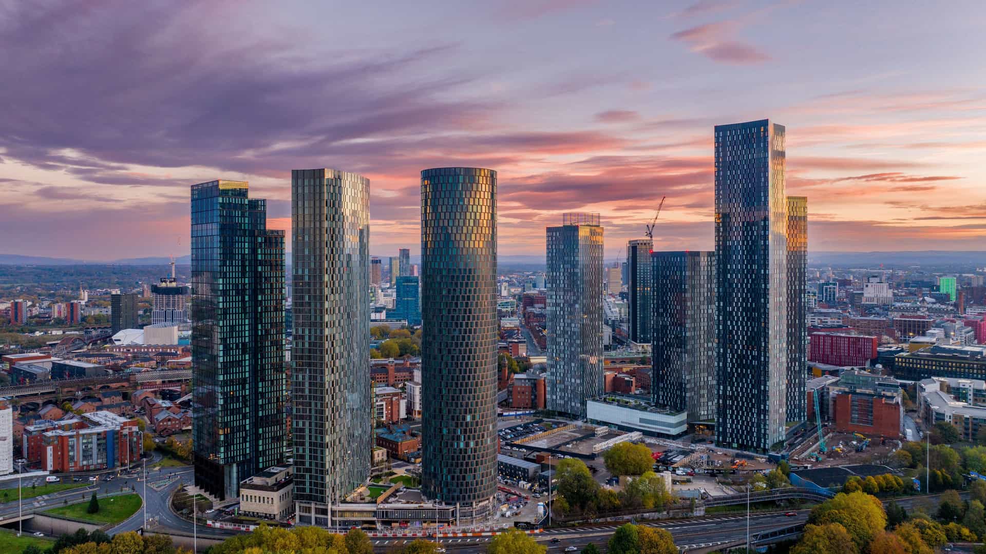 "An aerial sunset view of the modern residential skyscrapers of Deansgate Square in Manchester, England, reflecting the vibrant pink and orange clouds. "