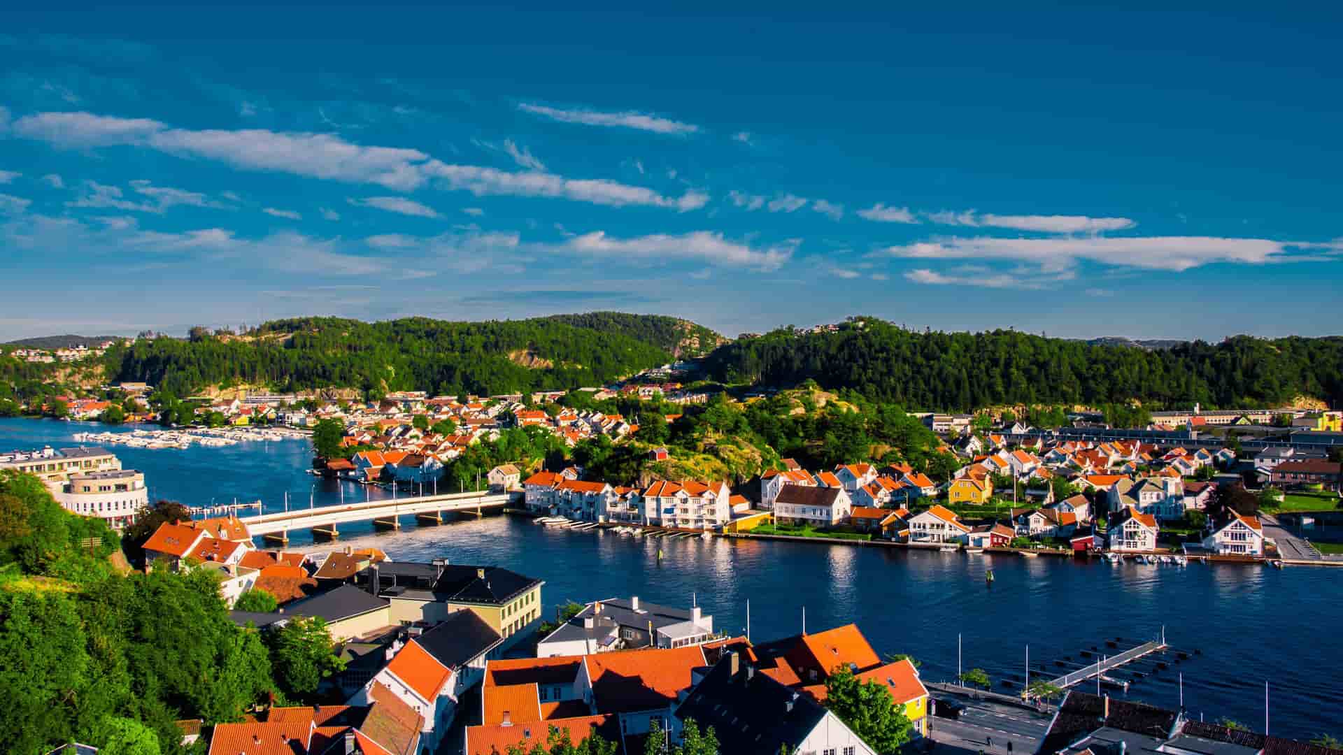 A scenic aerial shot of the coastal town of Mandal, Norway, with colorful houses clustered along the water and a bridge connecting two sides of the harbor.