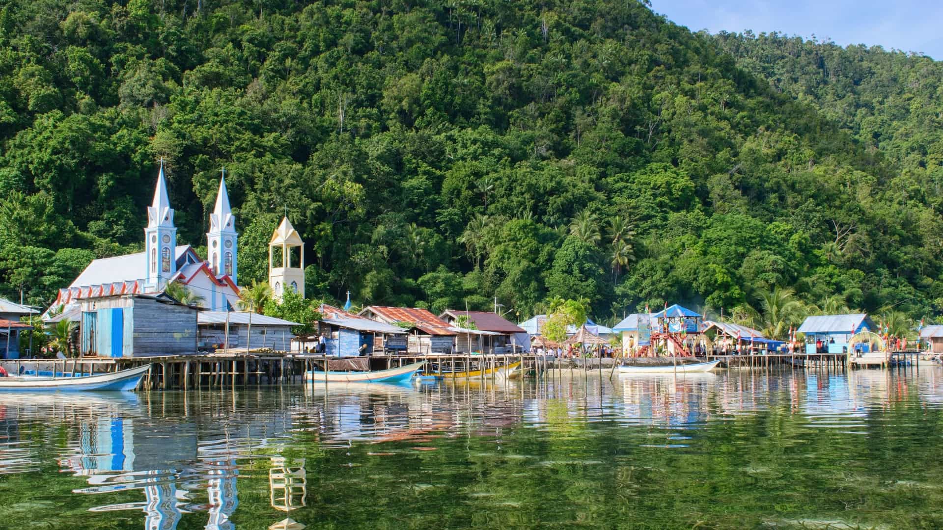 A waterfront view of a village on Mansuar Island, Indonesia, with wooden houses and a church on stilts, all nestled against a backdrop of a dense rainforest.