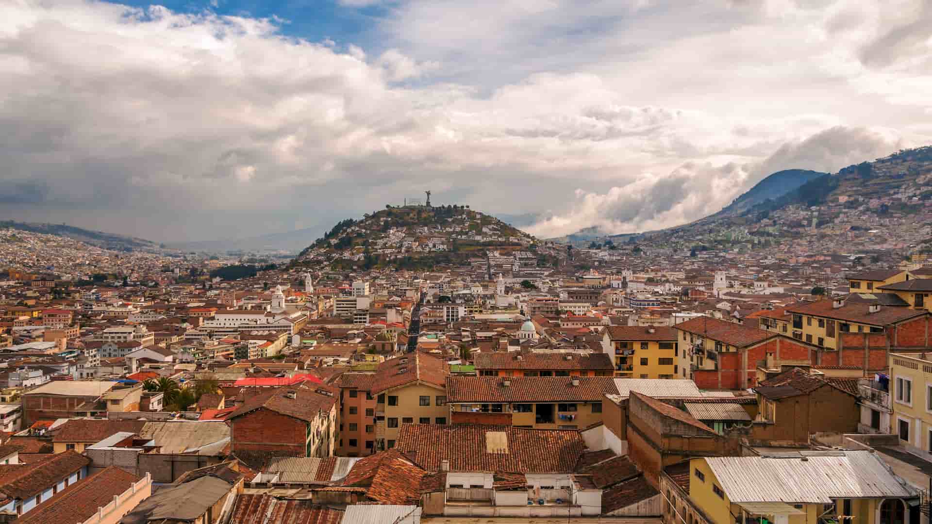 A scenic panorama of the coastal city of Manta, Ecuador, with a densely packed urban area surrounded by lush green hills under a dramatic cloudy sky.