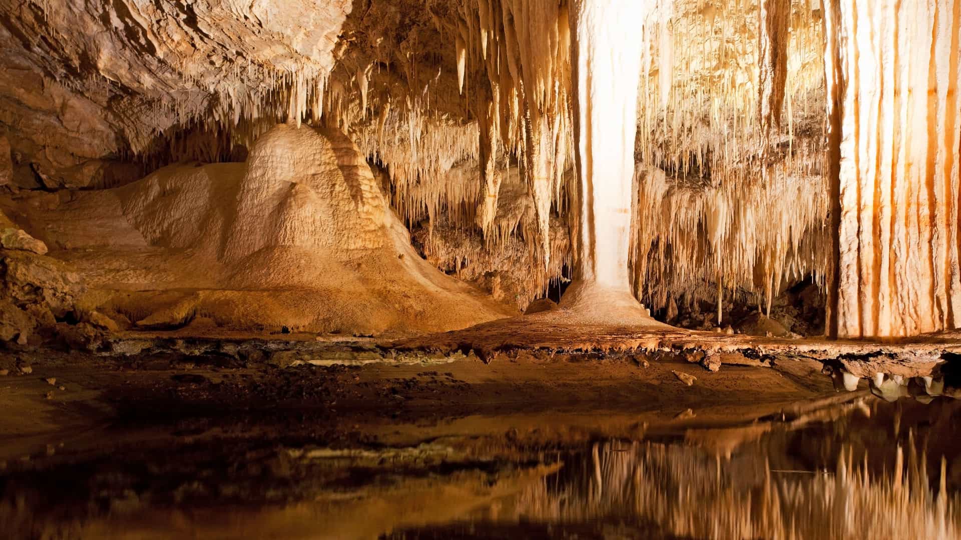 A dramatic view inside the Lake Cave in Margaret River, Australia, with a large "suspended table" formation and numerous stalactites reflecting in the still water below.